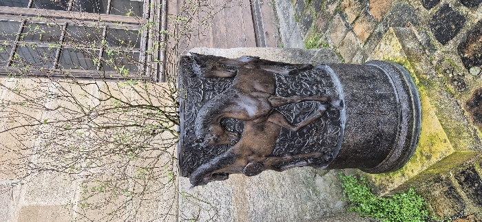 A cylindrical stone artifact featuring a relief carving of galloping horses dominates the scene. It sits atop a stone surface with mossy patches and is flanked by textured brickwork. A window with multiple panes is visible in the background, partially obscured by leafless branches. The carving details on the cylinder suggest a historical or artistic significance.
