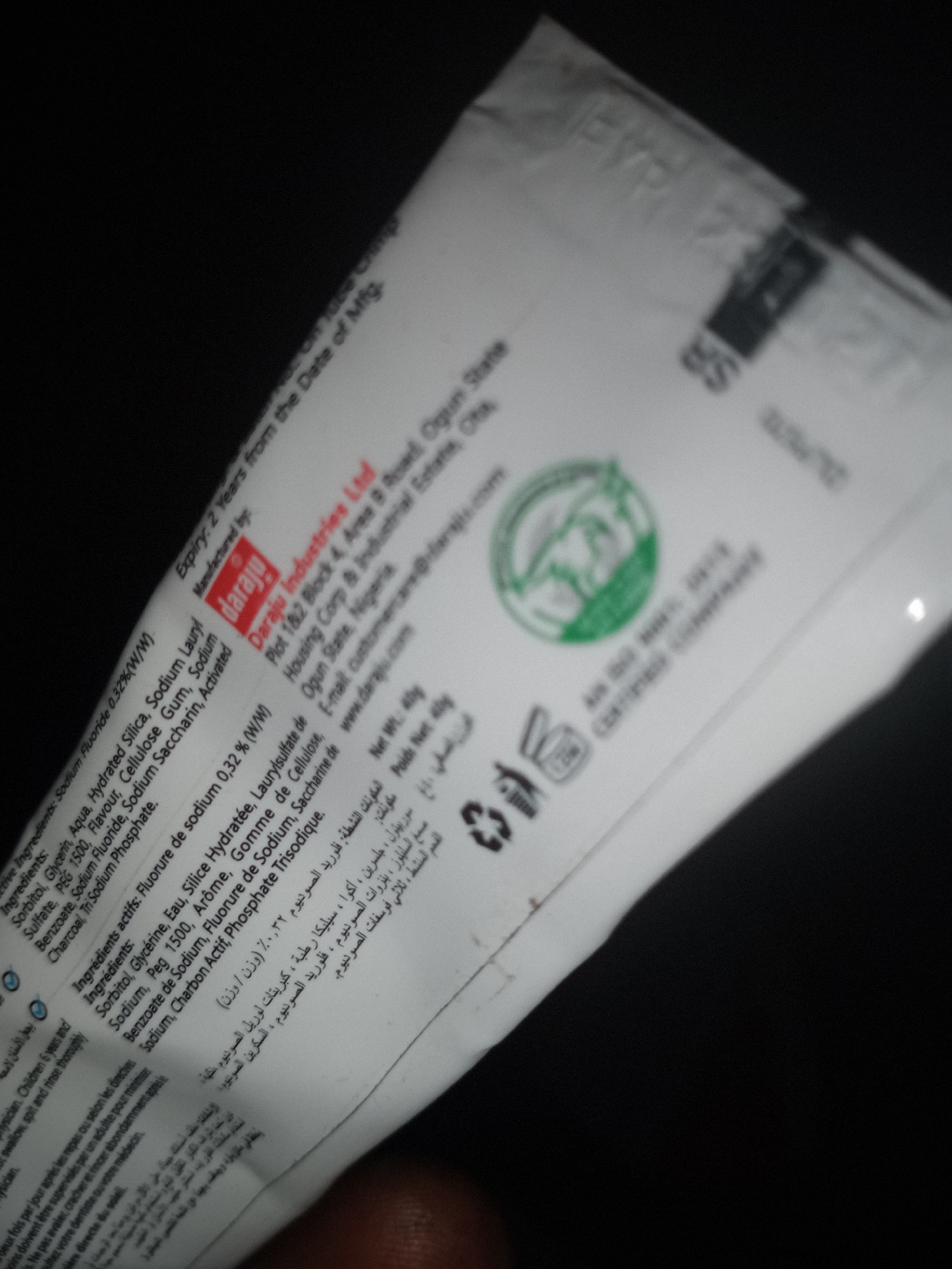 The image shows a close-up, slightly blurry, of a white tube, likely a toothpaste tube, with text and branding in various languages. The tube is angled, and the background is dark, suggesting it was taken in low light conditions. Text visible includes "Daraju Industries Ltd," "Daraju," and ingredient lists in English, French, and Arabic. There are also symbols indicating recycling and a duration of use after opening (12M). The overall impression is that of a product package. The image's quality does not provide sufficient detail to determine the specific location within Jalingo, Nigeria, or any people or actions occurring.