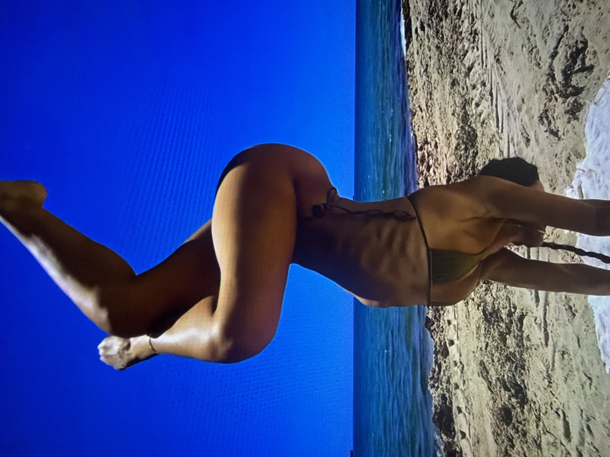 The provided image depicts an individual performing a handstand on a sandy beach, located in the city of Varna, Bulgaria. The person is inverted, with their upper body supported by their hands on a small white mat or towel placed on the sand. Their dark-colored hair appears to hang towards the ground. The individual is wearing a dark-colored bikini bottom, and a subtle dark marking or tattoo is visible on the left side of their torso. Their legs are bent at the knees, with the lower legs and feet curled upwards, positioned against a vibrant, cloudless blue sky. A thin, dark ankle bracelet is visible on the left ankle. The beach consists of light brown sand with scattered darker patches and small rocks, transitioning into a calm, blue-green ocean. A distinct horizon line separates the ocean from the sky, and an indistinct dark mass, possibly a ship or distant land, is visible on the horizon towards the left. The scene is brightly illuminated by natural sunlight, and the sky exhibits faint horizontal line artifacts consistent with digital capture or display.