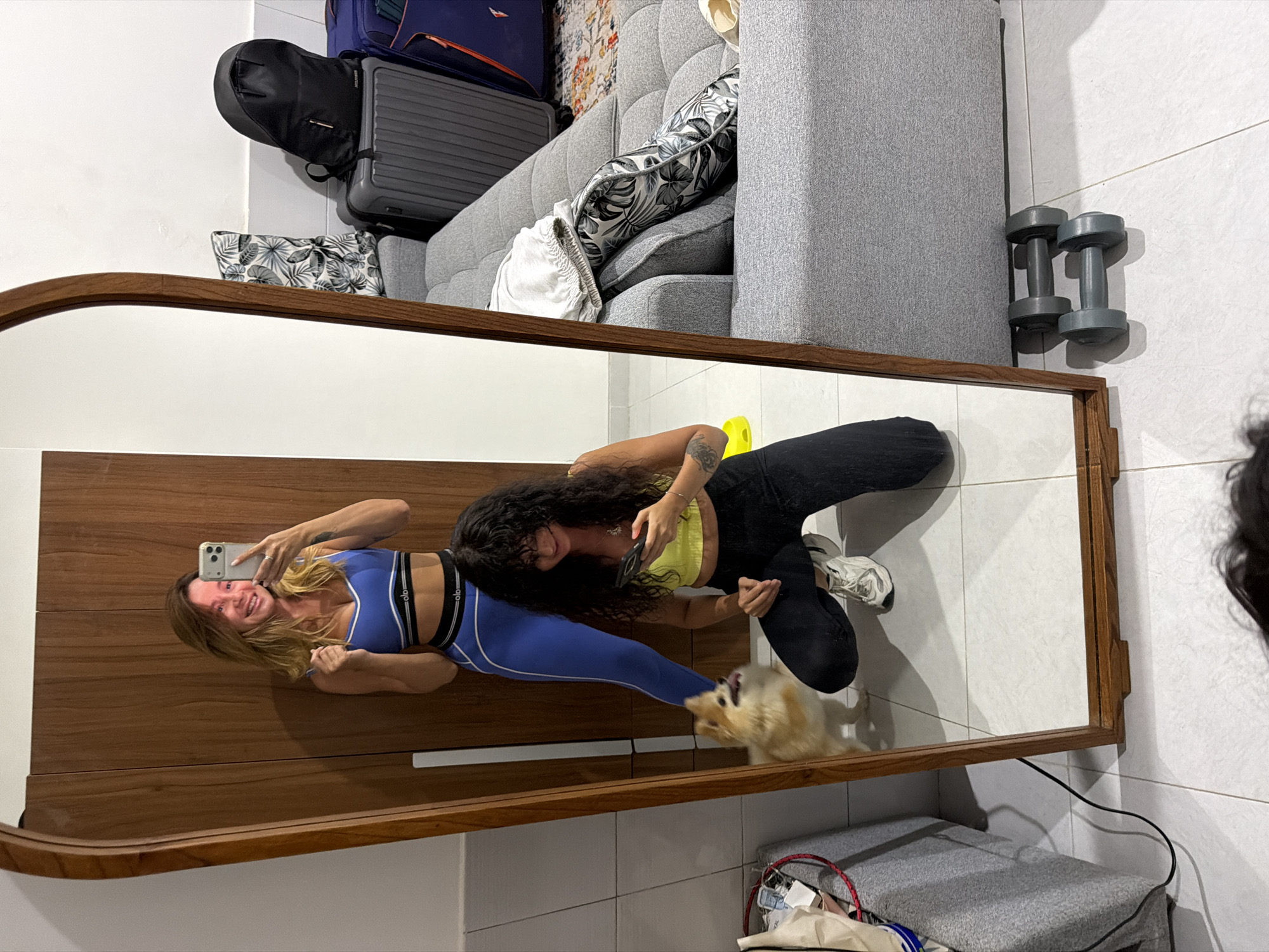 A full-length mirror reflects two women and a small dog in a room. The woman on the left, in the reflection, is taking a selfie with her phone. She has blonde hair and is wearing a blue sports bra and leggings. The woman on the right, also in the reflection, is looking at her phone and is wearing a yellow tank top and black pants. She is in a crouching position. A small, fluffy dog is standing between them. The room appears to be a bedroom or dressing area, with a wooden wardrobe, a grey sofa, luggage, and dumbbells visible in the reflection. The floor is tiled.