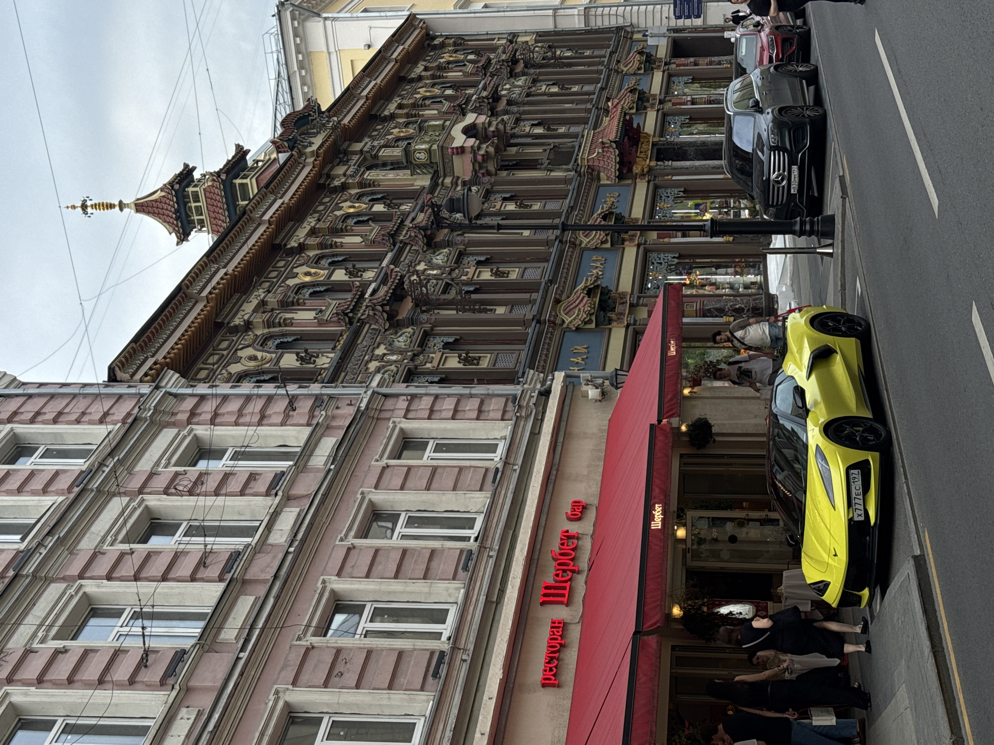 The image captures a street scene in Moscow, Russia, likely on a cloudy day. In the foreground, a bright yellow Chevrolet Corvette Stingray is parked along the curb. The building on the left has a pinkish facade with multiple windows and a red awning over a restaurant or bar called "Sherbet". The building on the right is elaborately decorated with intricate carvings and a distinctive tiered, pagoda-like rooftop structure, suggesting it is an older, architecturally significant building. Several people are visible on the sidewalk and near the restaurant entrance. A dark Mercedes-Benz GLS and a red SUV are parked behind the Corvette. The street is paved and marked with white lines.