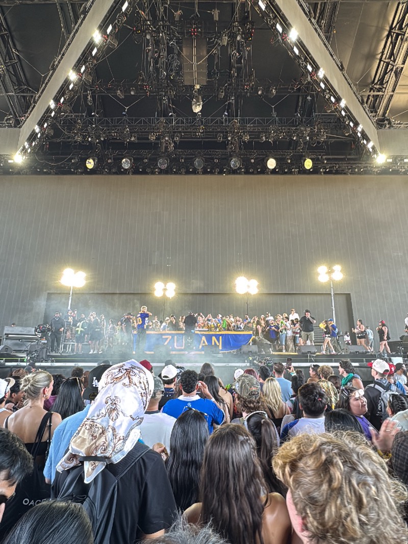 The image shows a crowd of people at a live performance in Palm Desert, United States. The concert is taking place on a large stage under a structure with lighting equipment and disco balls. A group of performers are visible on the stage, with the word "7UZUAN" displayed on a banner. The audience is densely packed, with various individuals visible wearing casual clothing and headwear.