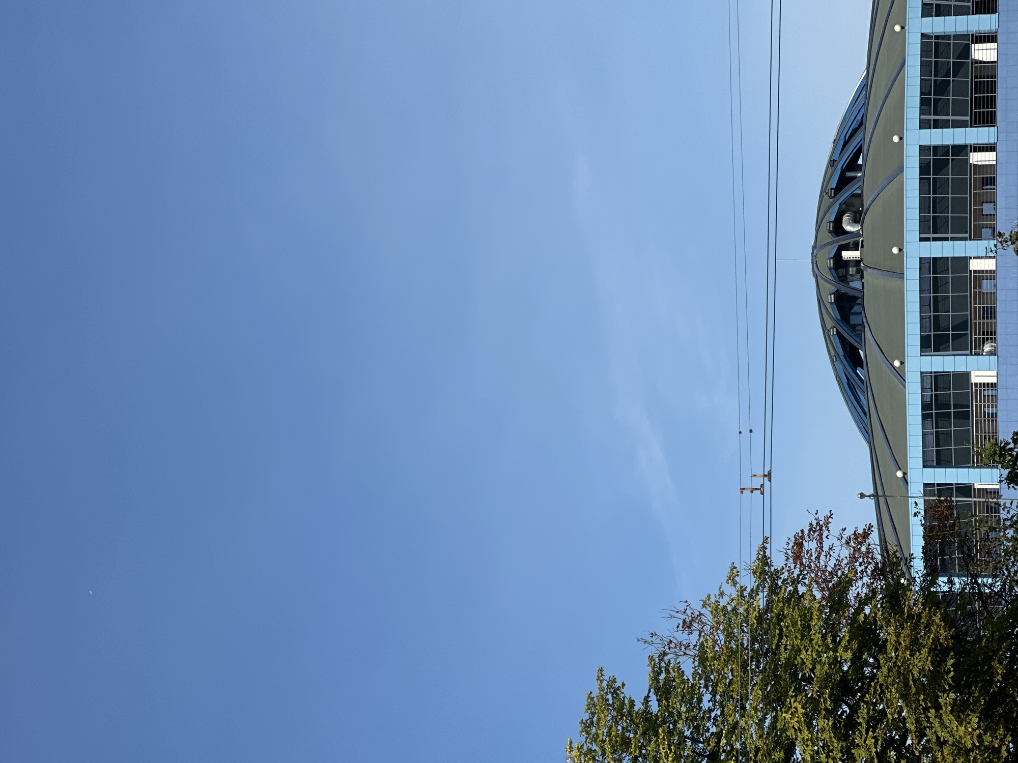 The image depicts the National Library of Belarus, a striking building known for its rhombicuboctahedron shape, situated in Minsk, Belarus. The photo is taken from a low angle, emphasizing the immense blue sky that dominates the upper portion of the frame. A crescent moon is faintly visible in the upper left quadrant of the sky.
The library's distinctive faceted roof, a dark green, is visible in the lower half of the image, contrasting with the bright blue facade of the building's lower levels. This facade features rows of large, grid-patterned windows. The roof is supported by a framework of dark struts, creating a geometric pattern. Several spherical lights are positioned along the edge of the roofline.
To the left of the library, a large tree with lush green foliage partially obscures the view, its branches reaching towards the sky. The leaves are vibrant, suggesting a warm season. The building is topped with what appear to be communication wires stretching horizontally across the sky, supported by small, red and brown poles. These wires add a layer of human infrastructure to the otherwise natural and architectural elements.
The overall impression is one of a clear, sunny day with a vast expanse of blue sky serving as a backdrop to the modern architectural marvel of the National Library. The composition highlights the building's unique design against the natural elements of the sky and the tree.