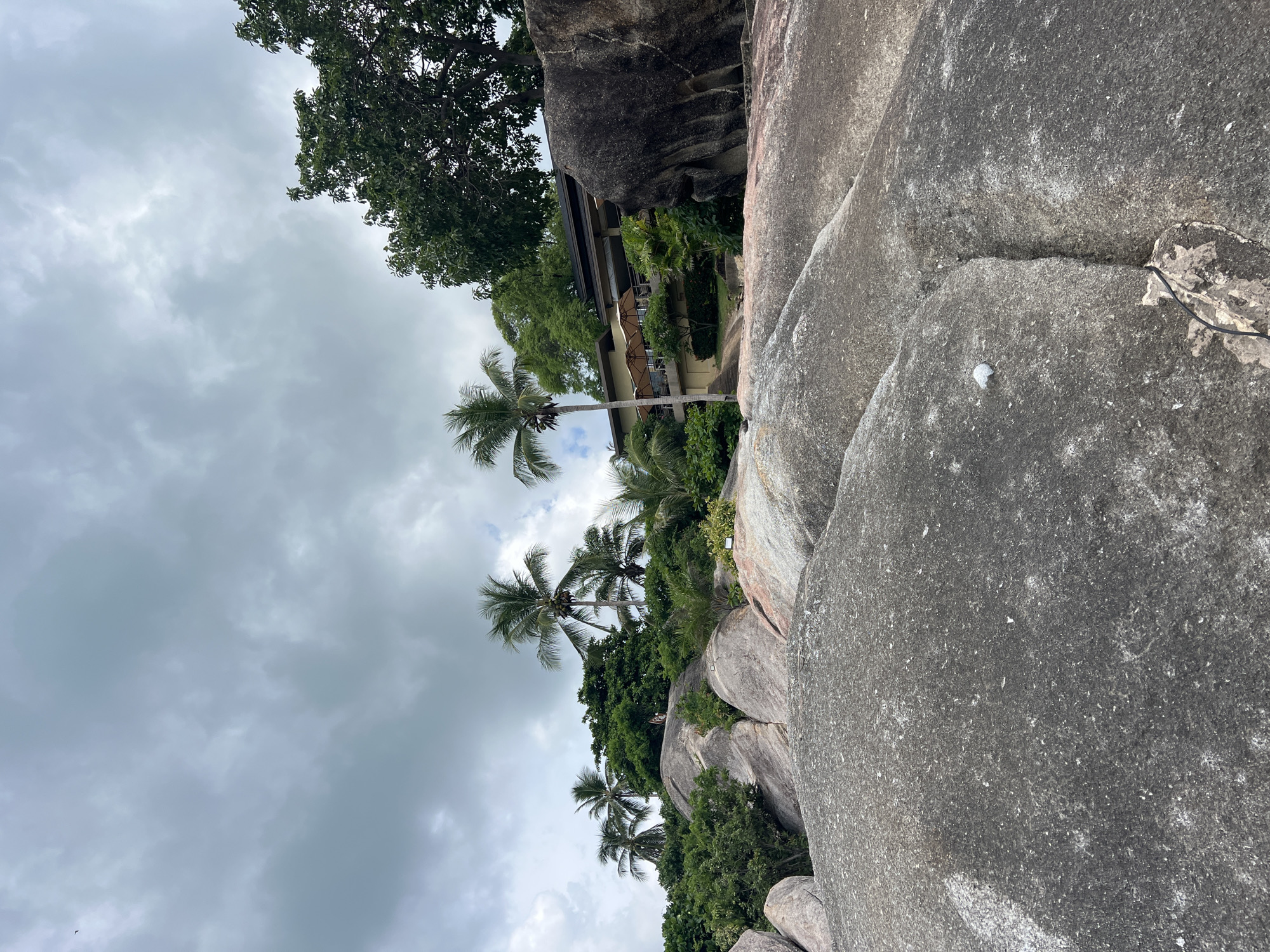 The image depicts a rocky coastal landscape in Baan Chaweng Noi, Thailand, under an overcast sky. The foreground is dominated by large, smooth grey boulders, some showing pinkish hues. Beyond the boulders, lush green vegetation, including palm trees, covers a hill. A building with a light-colored facade and a dark roof is visible nestled amongst the trees on the hill. The building has a balcony with several large, light-brown umbrellas. A single person can be seen standing on the rocks near the vegetation, facing away from the camera. The environment appears natural and tropical, with a sense of serenity.