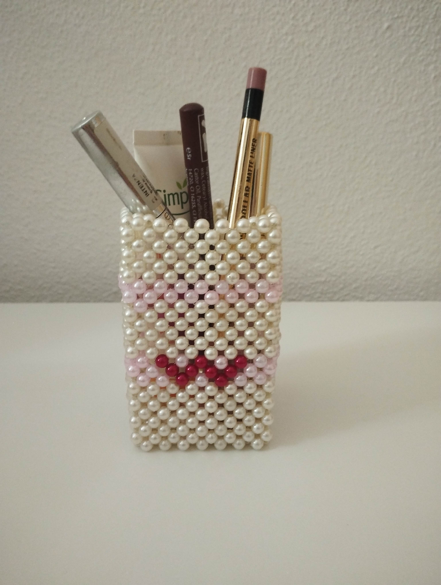 An eye-level shot captures a rectangular container handcrafted from small, pearl-like beads, situated on a bright white surface against a light, textured wall. The container is predominantly made of off-white beads, with a playful pattern resembling a stylized face or a heart formed on its front using lighter pink and darker red beads.
Inside the beaded holder, several cosmetic items are stored upright. These include a silver-toned tube with partial text "INTENZA" visible. Next to it is a white tube, showing part of a brand name "imp" and ingredient details such as "Castor Oil". A dark brown pencil or crayon is also present, marked with "esr" and further ingredient information like "wax, Cetearyl Alcohol". Another item is a golden pencil, clearly labeled "OLLAR MATTE LINER" (likely "DOLLAR MATTE LINER") on its shaft, topped with a pinkish-brown tip. A second, less distinct gold-colored item stands beside it.
The scene is well-lit, suggesting an indoor setting, possibly during the daytime or under bright artificial light. The image solely focuses on these static objects, showcasing them in an organized manner.