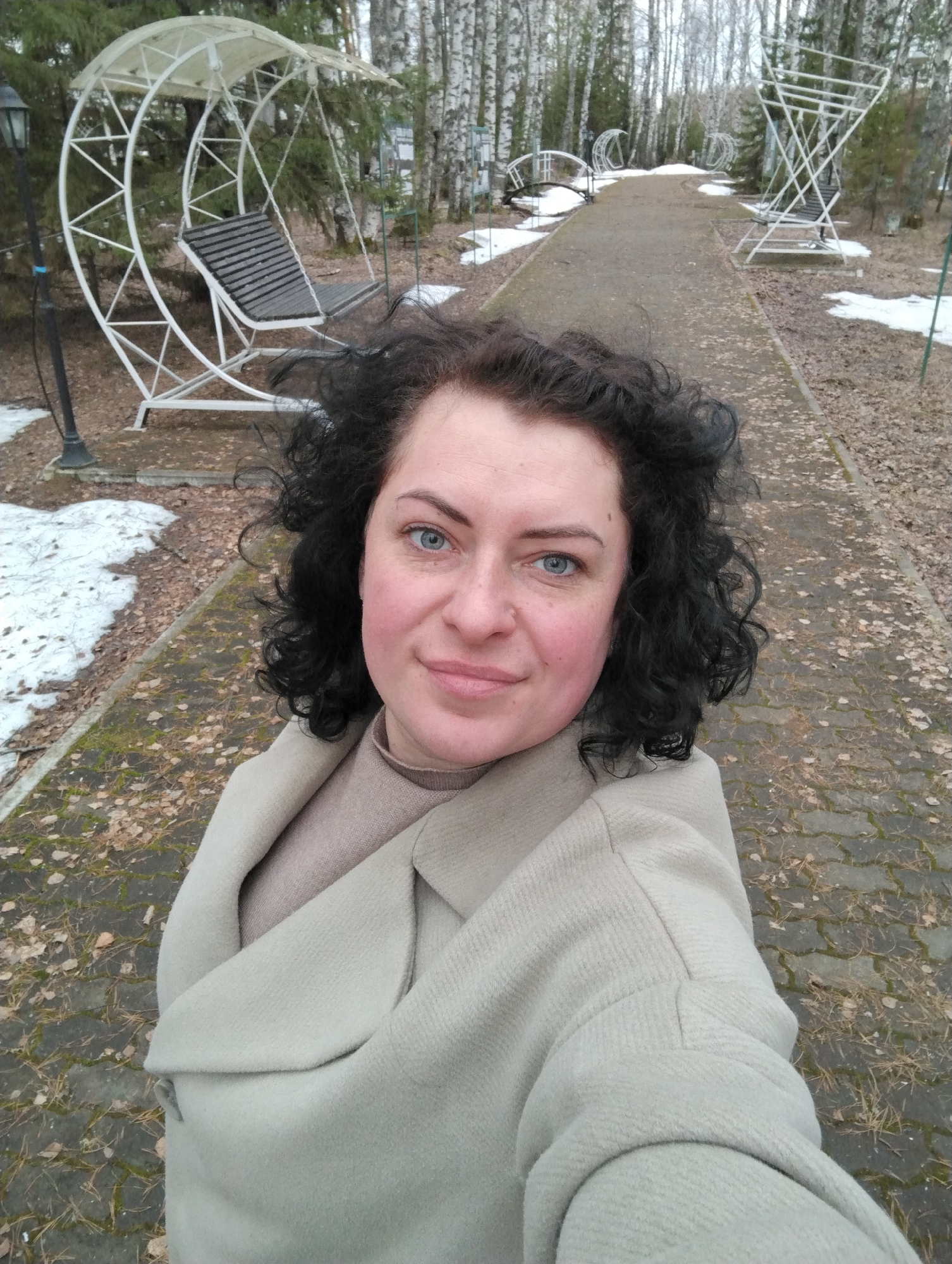 A middle-aged woman with fair skin, blue eyes, and short, curly black hair takes a selfie while outdoors. She is wearing a tan overcoat and a light brown turtleneck sweater. The background shows a path paved with patterned grey stones, covered with dry leaves. Along the path are white metal swing sets, and patches of melting snow. Bare trees line the path, indicating it is late fall or early spring. The image was taken in Galevo, Russia.