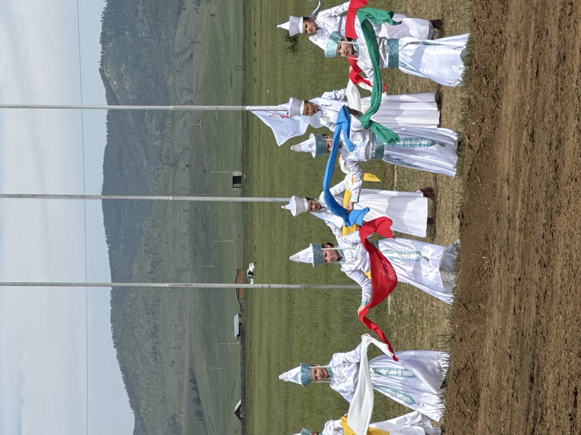 In Orongoi, Russia, a group of women in traditional white and blue attire, complete with pointed hats, perform a dance in a grassy field. They are holding colorful silk scarves, red, white, green, and blue, which they manipulate in unison. The background features rolling hills covered in green vegetation and scattered trees, with several tall poles, one bearing a white flag with red markings, standing in the middle ground. In the distance, a few wooden buildings and a white vehicle are visible. The foreground consists of a patch of brown, disturbed earth.