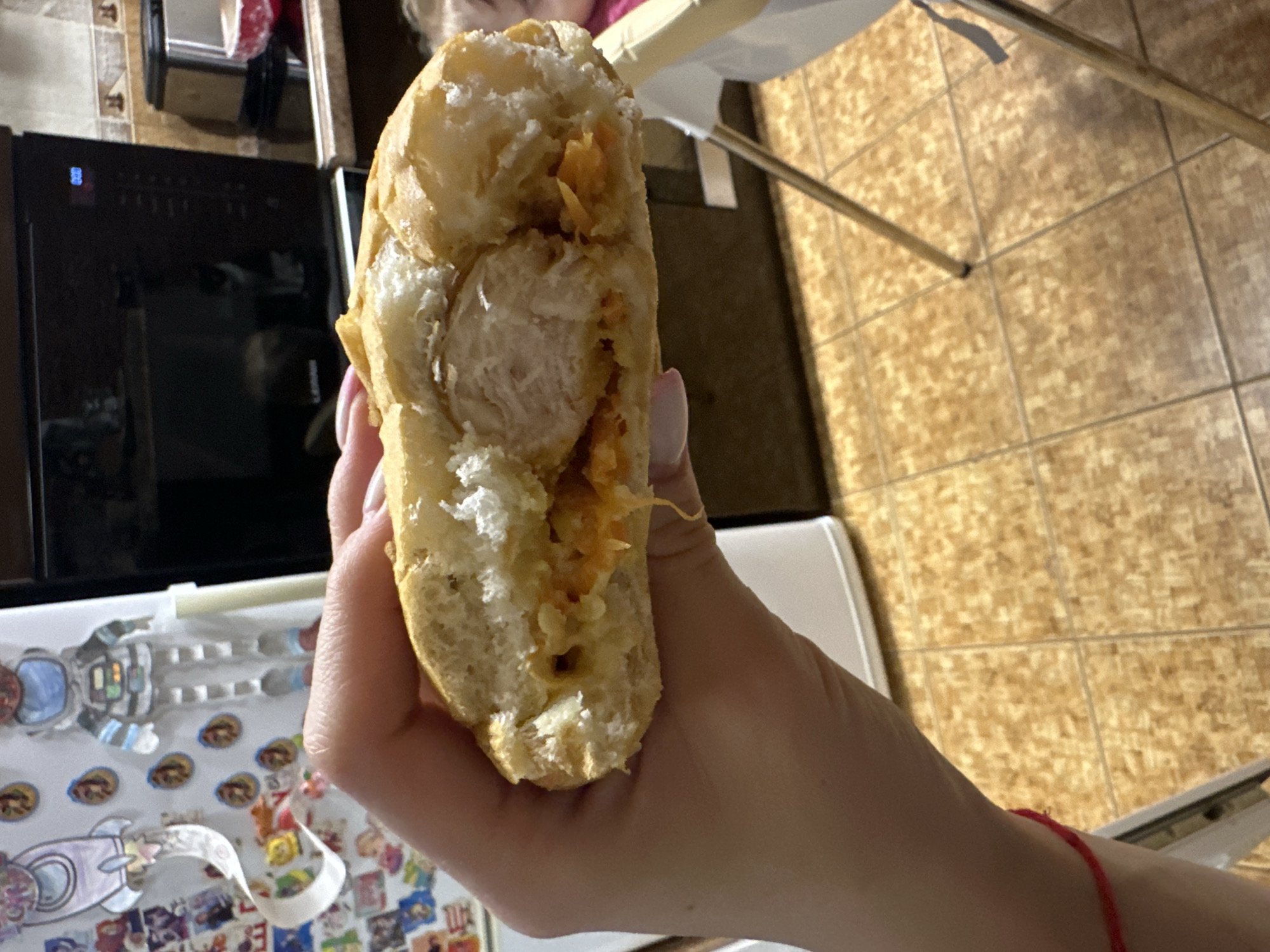 A person's hand holds a half-eaten sandwich over a tiled floor. The sandwich has a golden-brown bun and is filled with a piece of white meat and a shredded orange substance, possibly shredded carrots or cheese. The background shows a kitchen with a microwave and refrigerator. A red string is tied around the person's wrist. The scene does not provide any information about the city of Sayanogorsk, Russia.