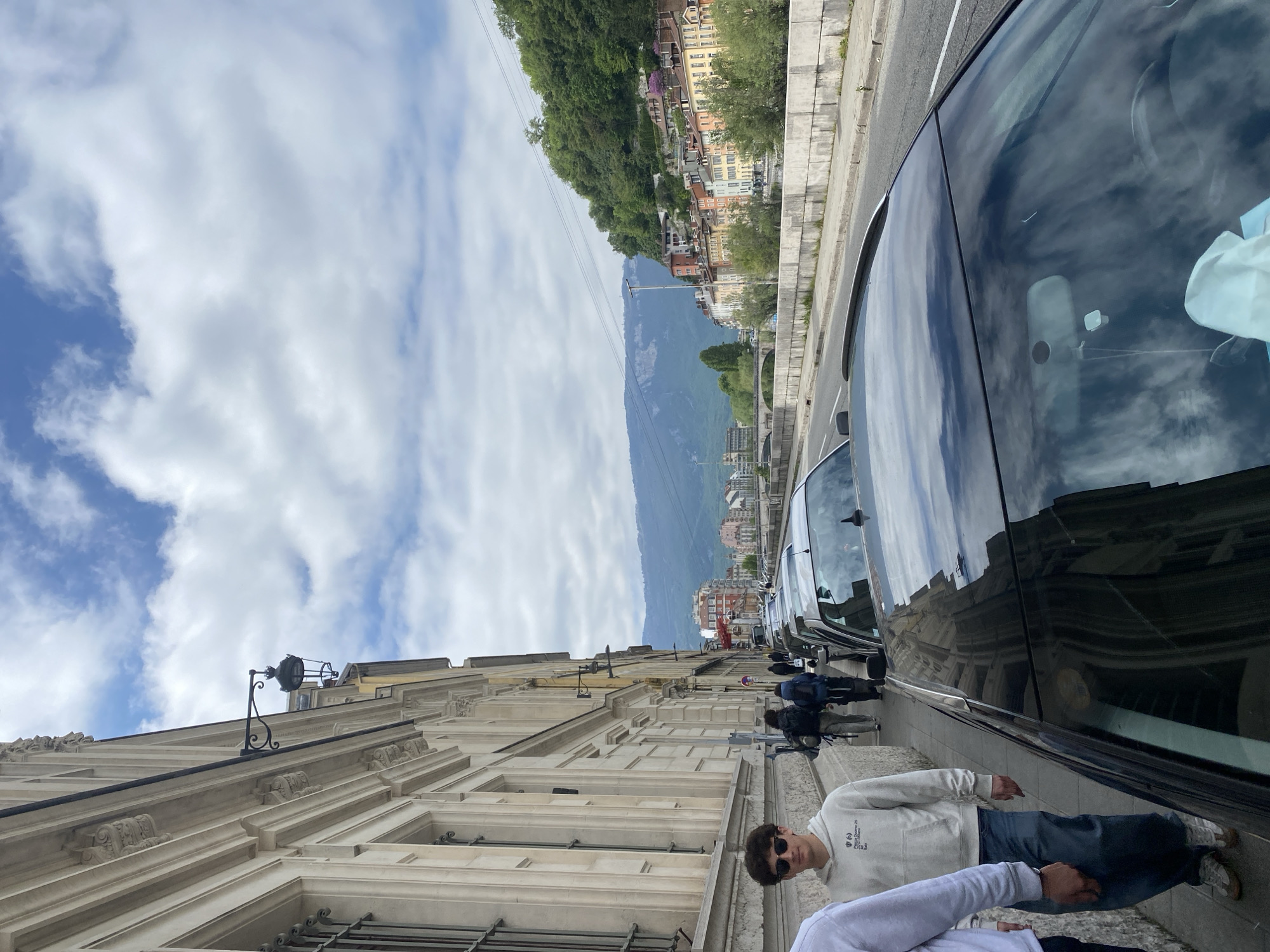 Here's a description of the image:
The image presents a street scene. The photo is taken from the sidewalk, facing a street lined with vehicles. On the left is a building with ornate architectural details. People are walking on the sidewalk. In the background, across the street, is a hillside with buildings. Mountains are visible in the distance. The sky is cloudy. The location appears to be in a European city. Grenoble, France is the city.