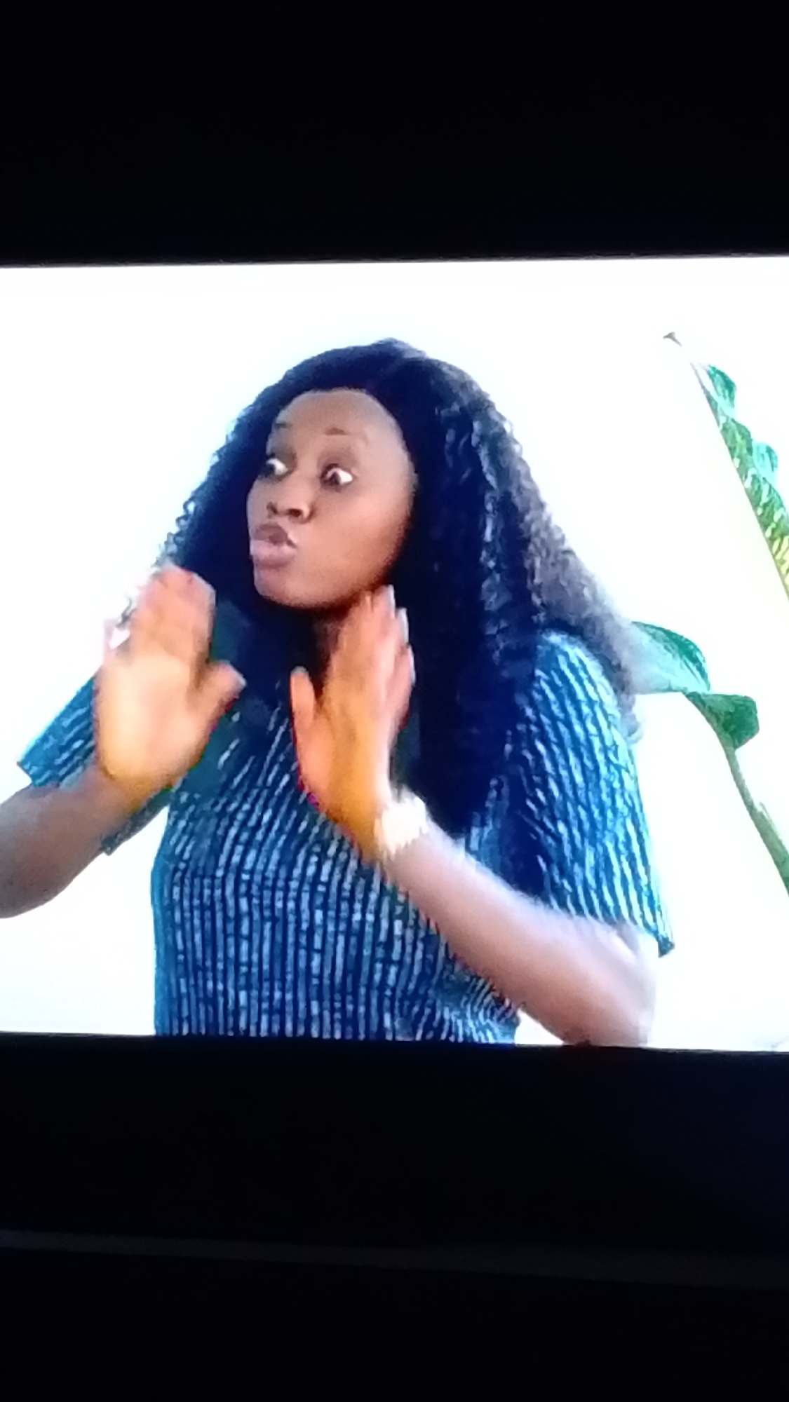 A close-up shot captures a Black woman from the chest up, against a plain white backdrop. She has dark, curly hair and is wearing a blue and white striped shirt. Her head is turned to her left, and her mouth is slightly open, giving the impression she is speaking or making a sound. Her hands are raised in front of her, palms facing outwards, in a gesture that could be interpreted as warding off or emphasizing a point. A watch is visible on her left wrist. The overall scene is brightly lit, with a hint of a green plant on the right side of the frame. There is no visible indication of the city of Lokoja, Nigeria.