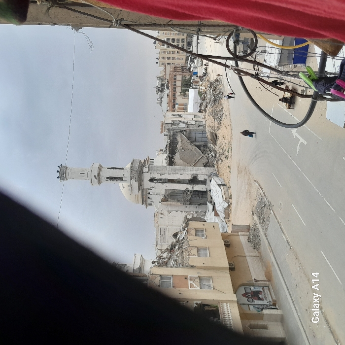 This image depicts a scene of widespread urban destruction in Khan Yunis, Palestinian Territory, under an overcast sky. The focal point is the severely damaged Great Mosque of Khan Yunis (Al-Kabir Mosque), centrally positioned. Its distinctive minaret shows visible scarring and structural compromise, while large sections of the main building and dome have collapsed, exposing internal frameworks and scattering significant piles of rubble around its base.
To the left of the mosque, a multi-story building exhibits extensive damage, including a partially collapsed roof and visible impact marks on its facade. On the lower part of this building's facade, faded graffiti features symbols from 'Squid Game' alongside Arabic text.
A wide, paved road with white lane markings occupies the foreground and middle ground, leading towards the damaged structures. A lone individual is observed walking on this road, moving away from the viewer, carrying an item. Further to the right on the road, several people are seated or standing near a low structure. In the mid-distance, beyond areas of sandy ground and accumulated rubble, additional figures are visible in what appears to be a cleared area.
The photograph is taken from an elevated perspective, evidenced by structural elements, wires, and a red cloth partially framing the right side of the image. A dark, out-of-focus object partially obscures the upper-left corner. A "Galaxy A14" watermark is present in the lower-left corner.