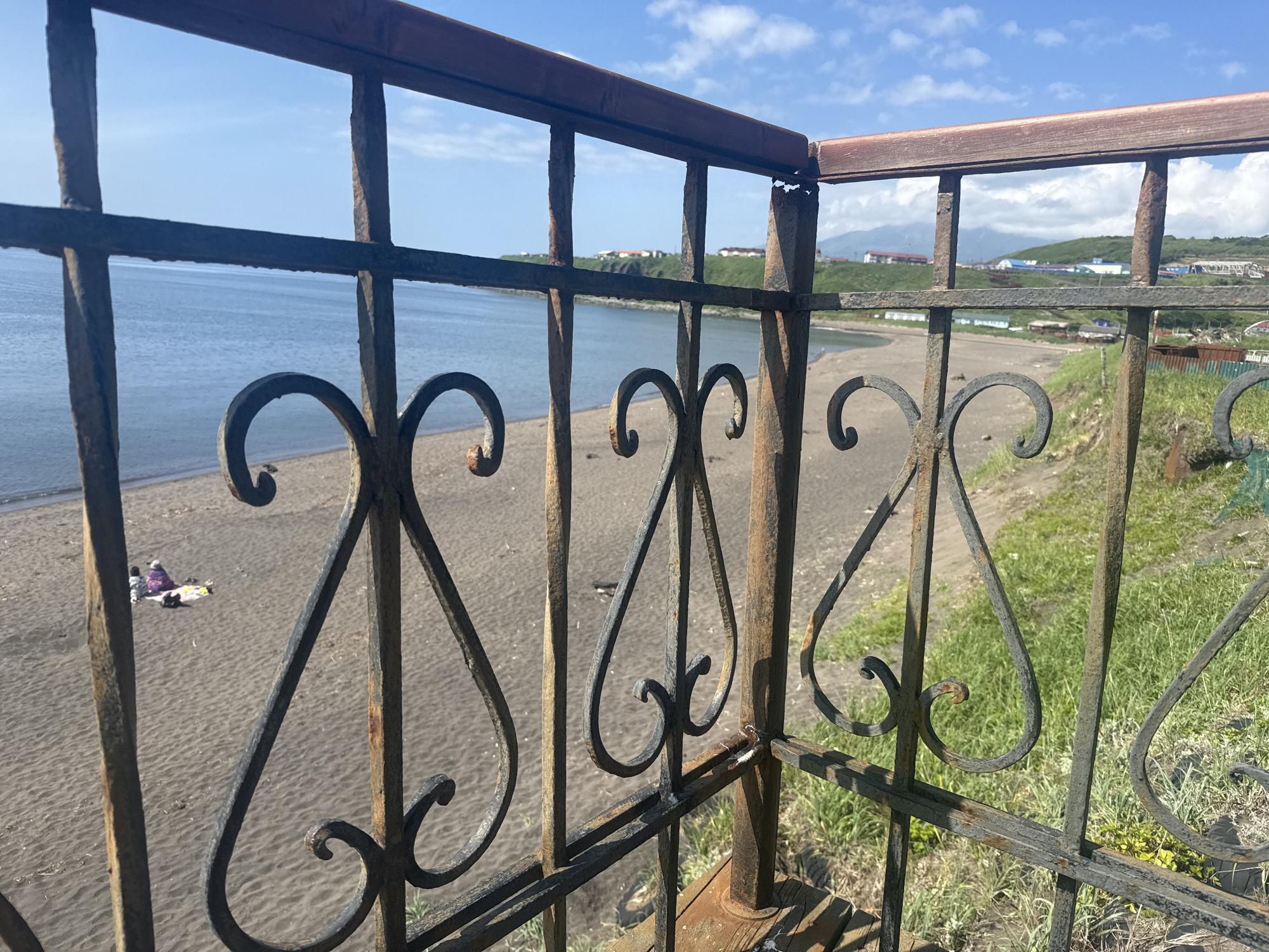 The image shows a view of a beach and the sea through a rusty metal fence. The beach appears to be dark sand. In the distance, there are some buildings on a hill overlooking the water. A small group of people are sitting on the beach on a blanket. The sky is blue with some white clouds. The fence has decorative elements that are made of metal. The overall impression is of a somewhat weathered and rustic coastal scene in Kuril'sk, Russia.