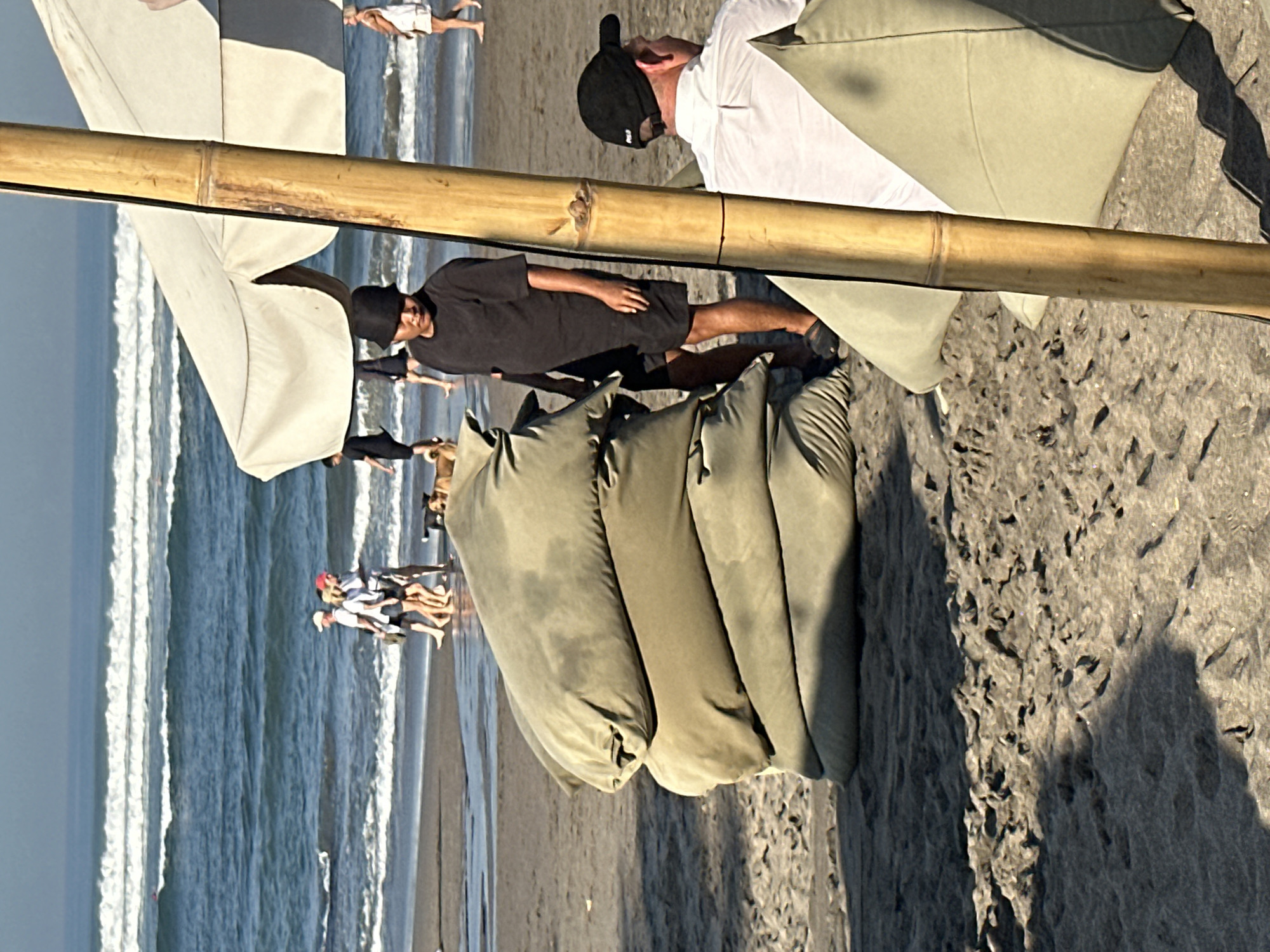 The image depicts a beach scene in North Kuta, Indonesia, likely in the late afternoon given the lighting. In the foreground, a stack of large, olive-green beanbag chairs sits on the sand. To the right of the beanbags, a tall, thick bamboo pole, part of a beach umbrella structure, is prominent. A man wearing a black bucket hat and a dark t-shirt and shorts stands behind the beanbag chairs, facing away from the camera. To the right and slightly behind him, another man wearing a white shirt and a black baseball cap is seated, facing to his left. In the background, the ocean with gentle waves is visible, and several people are walking along the shoreline, some with dogs. A white beach umbrella is partially visible in the upper right of the frame. The overall atmosphere suggests a relaxed beach environment.