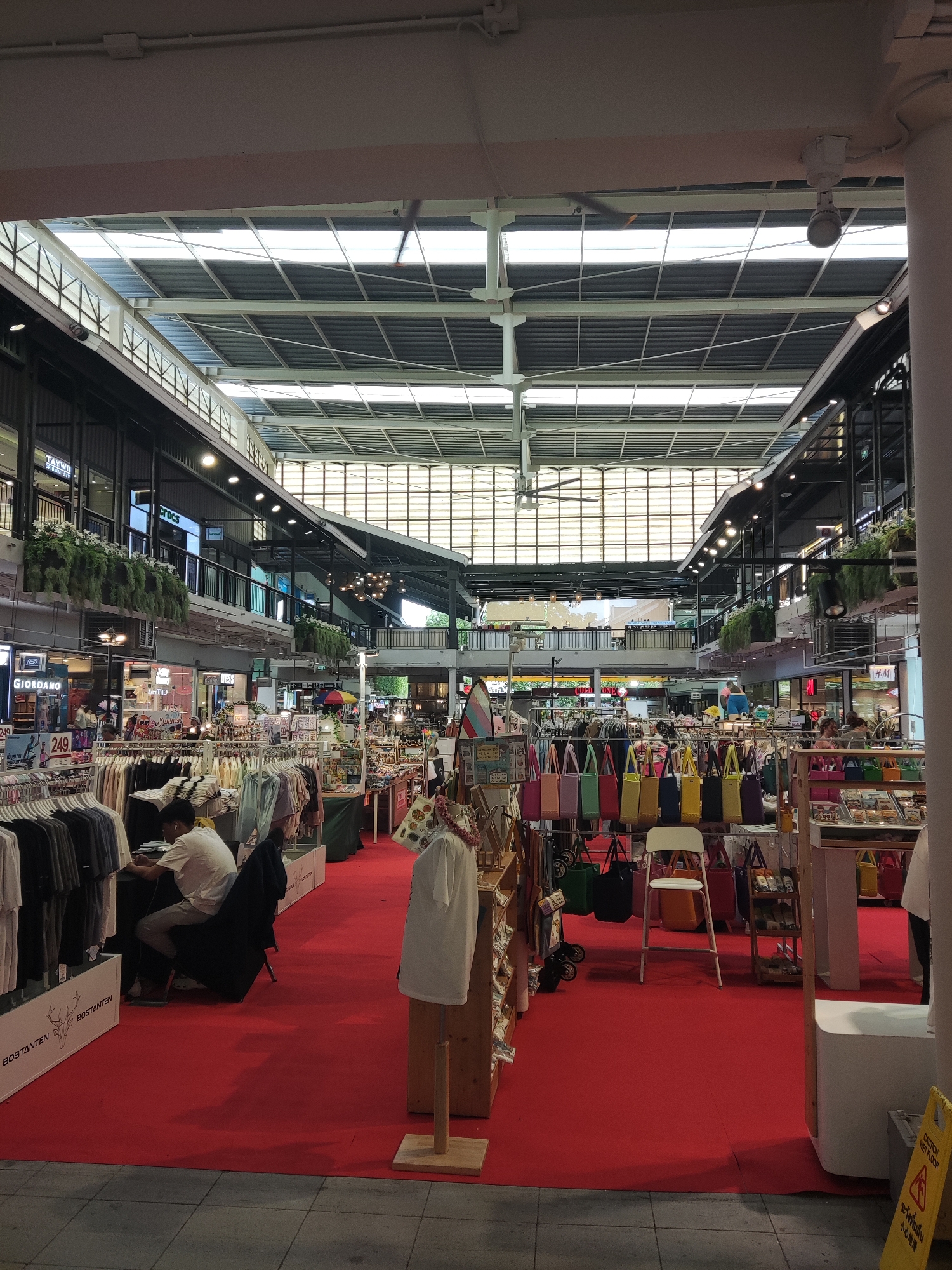 The image depicts a bustling indoor marketplace or shopping center in บ้านบางรักษ์, Thailand. The space is characterized by a high, multi-tiered ceiling with a skylight design, allowing natural light to illuminate the area. Shops and stalls line the perimeter, featuring various merchandise, including clothing, bags, and miscellaneous goods. People are scattered throughout the scene, some browsing the items on display, while others seem to be working at the stalls. A large red carpet covers a significant portion of the floor, possibly delineating specific vendor areas. Signage and brand names are visible on storefronts and displays.