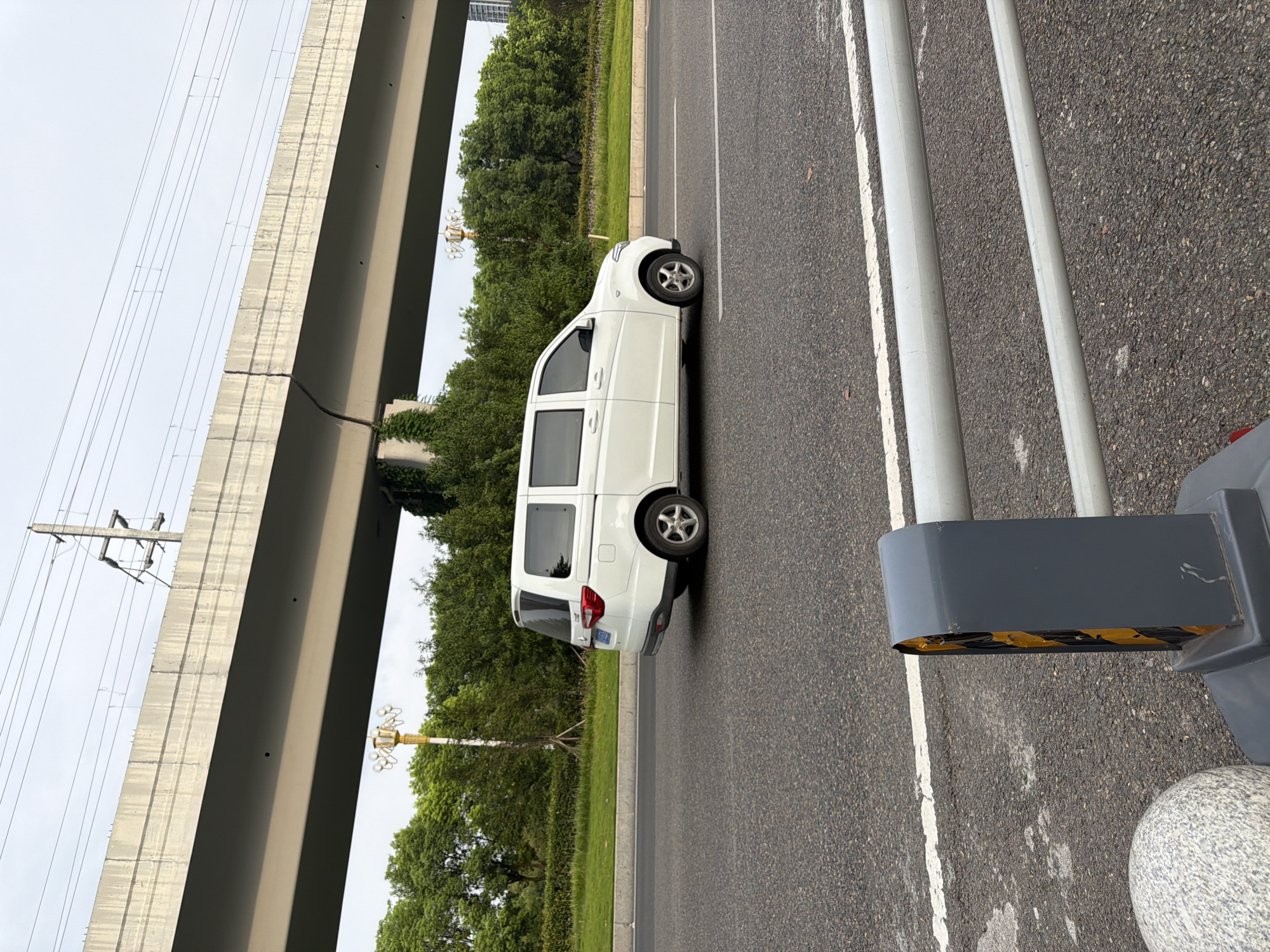 A white minivan is captured in profile, traveling on an asphalt road. The vehicle is positioned on the right side of the road, moving from left to right within the frame. Behind the minivan, a lush green landscape with trees and grass is visible. Overhead, a concrete overpass with power lines and supporting structures stretches across the scene. The sky is overcast. In the foreground, a grey barrier with yellow reflective tape and a rounded granite bollard are present. The image appears to be taken in Jiangjin, China, based on the context provided.