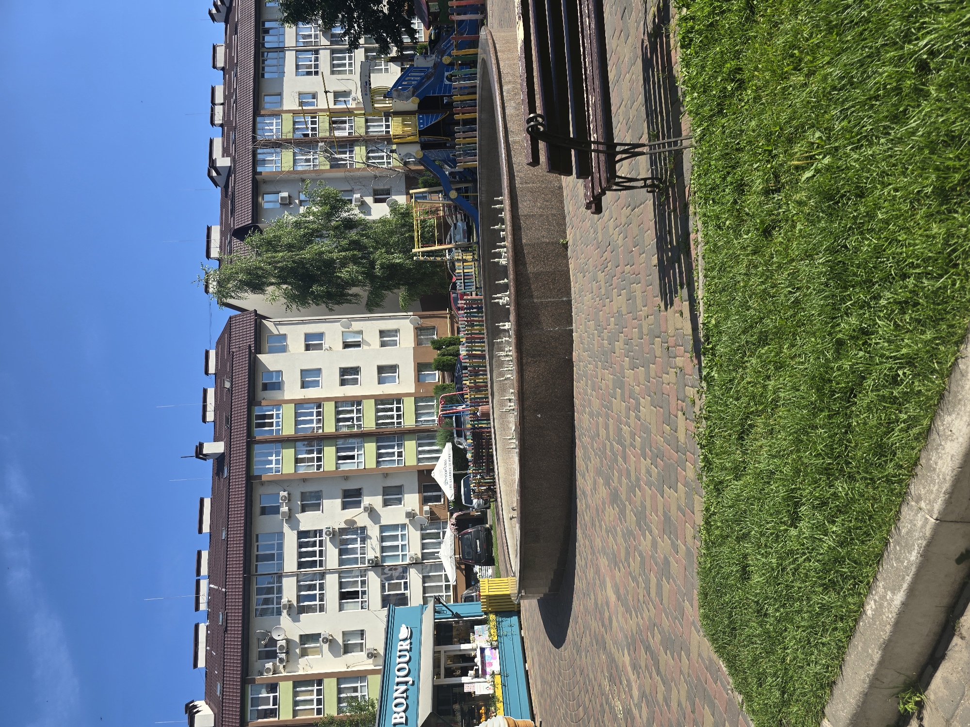 In Petropavlivska Borshchahivka, Ukraine, a residential courtyard is depicted under a clear blue sky. The scene features modern apartment buildings with beige and green facades, featuring numerous windows and air conditioning units. In the foreground, a circular stone fountain with a series of water spouts is centrally located. To the left of the fountain is a light blue kiosk with the word "BONJOUR" prominently displayed, suggesting a small business, possibly selling ice cream or refreshments. Adjacent to the kiosk, a patch of vibrant green grass meets a decorative paved walkway constructed from interlocking bricks in shades of red, yellow, and gray. A dark wooden park bench with wrought iron legs is positioned on the paved area, facing the fountain. Behind the fountain and to the right, a colorful children's playground is visible, complete with slides, climbing structures, and a protective fence. Several cars are parked near the buildings and playground. The overall environment is tidy and well-maintained, indicative of a residential area designed for communal use and recreation.