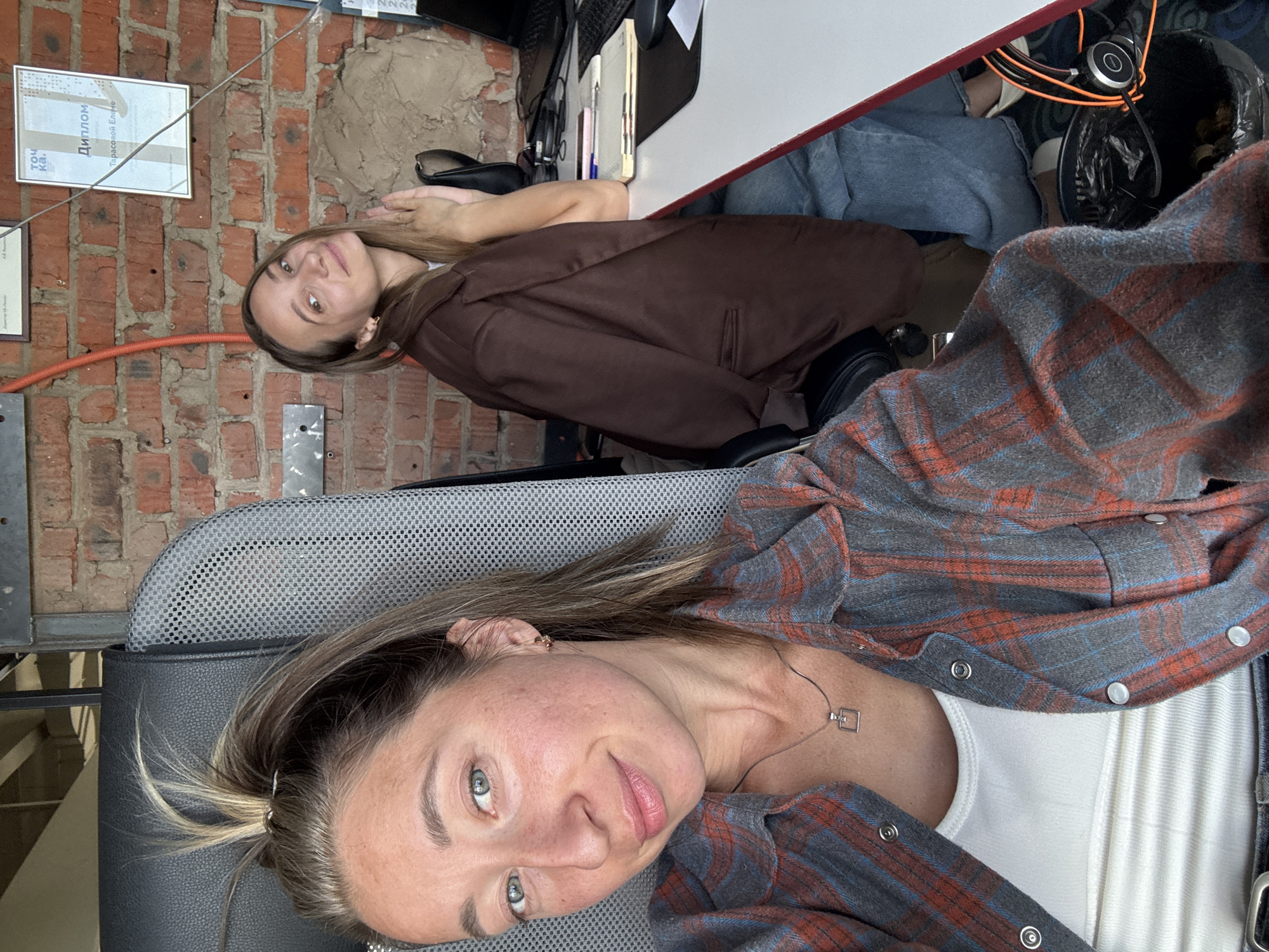 The image is an indoor photograph of two fair-skinned women. The woman closest to the camera is seated in a dark-colored office chair. She wears a white top covered by a red-and-grey plaid flannel shirt and a silver necklace with a square pendant. Her light brown hair is partially pulled back, and she is looking directly into the camera. Behind her, another woman with long brown hair and a brown blazer is seated at a desk. A certificate in Russian is on the brick wall. A desk with a computer and various items on it is near the wall. The location is Chaikovsky, Russia.