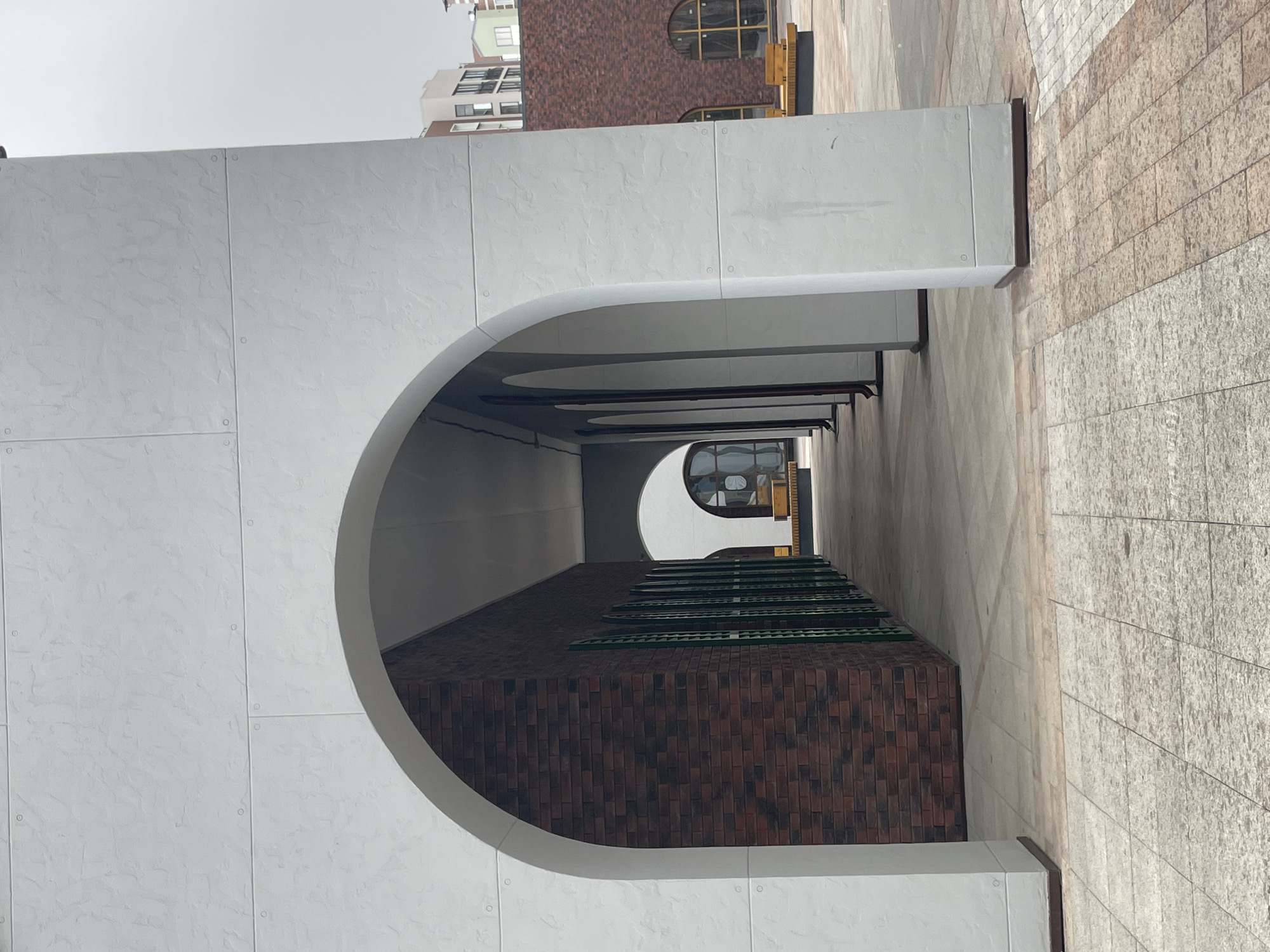 An outdoor architectural scene in Izhevsk, Russia, depicts a series of repeating archways forming a covered walkway or arcade. The main subject is a large, white archway in the foreground, constructed from panels with visible fasteners and a textured surface. Through this arch, a perspective view extends down a corridor created by similar receding arches.
To the right of the covered walkway, a building wall made of reddish-brown bricks is visible, topped with a series of long, green, slatted grates or vents. Further down the corridor, another brick building with distinct circular or arched windows can be seen. The ground is paved with light-colored, possibly granite or concrete, tiles arranged in a grid pattern.
In the upper background, above the white architectural elements, a modern, multi-story building with dark-framed windows is partially visible. The sky above is overcast, suggesting daytime with soft, diffused lighting, as there are no harsh shadows. No people or visible text are present in the image. The overall aesthetic is one of clean lines and geometric forms, blending modern design with traditional brickwork.