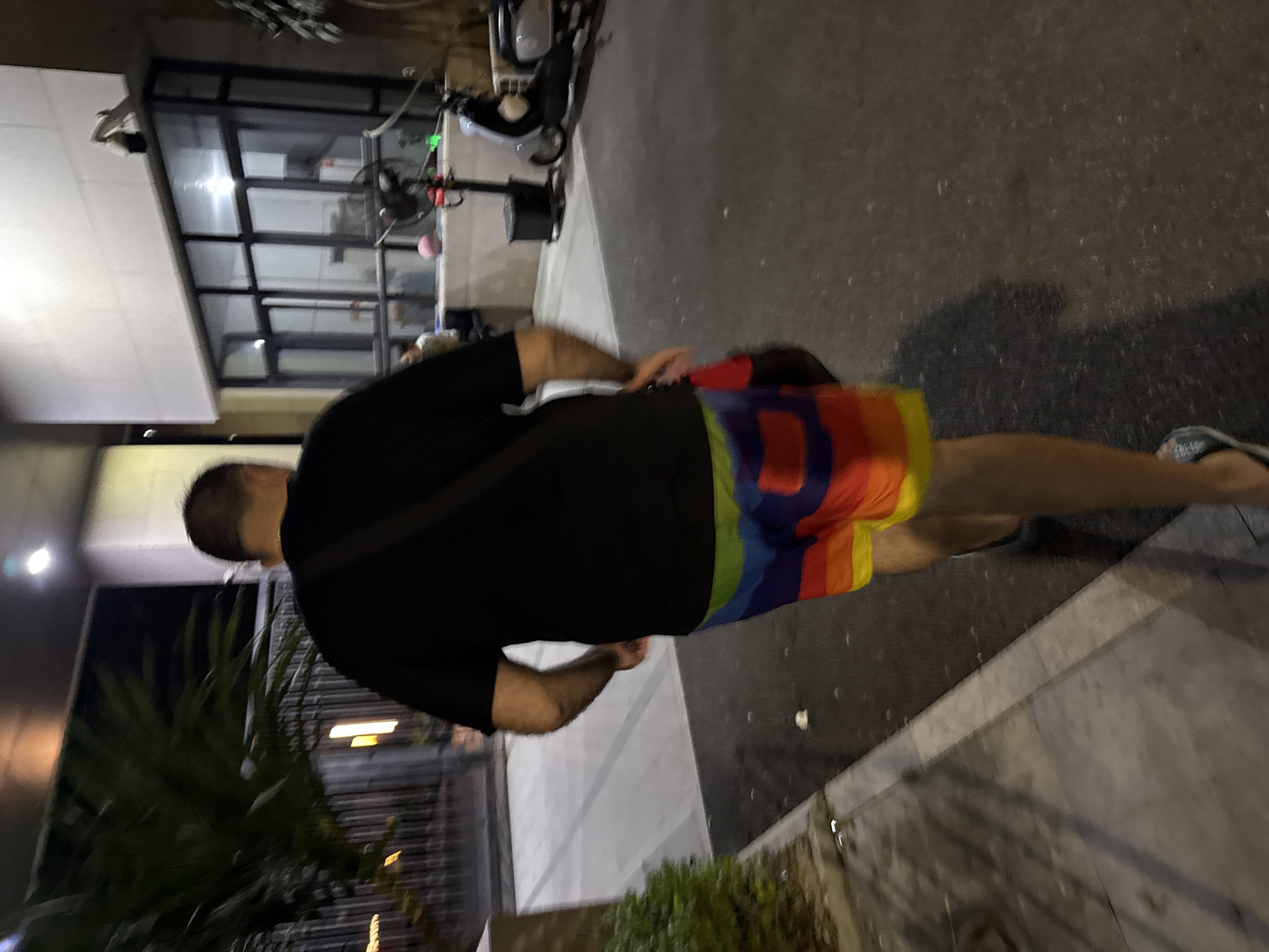 A medium shot captures a person from behind, walking away from the camera on a paved surface. The individual is wearing a black t-shirt and multi-colored rainbow-striped shorts. A dark strap crosses their chest. The environment suggests an urban setting at night, with a building featuring large windows to the right and a palm tree to the left. A security camera is visible on the building's exterior. A scooter is parked near the building. The ground is wet, indicating recent rain. The lighting is dim, with artificial lights illuminating parts of the scene. The location is identified as Huizhou, China.