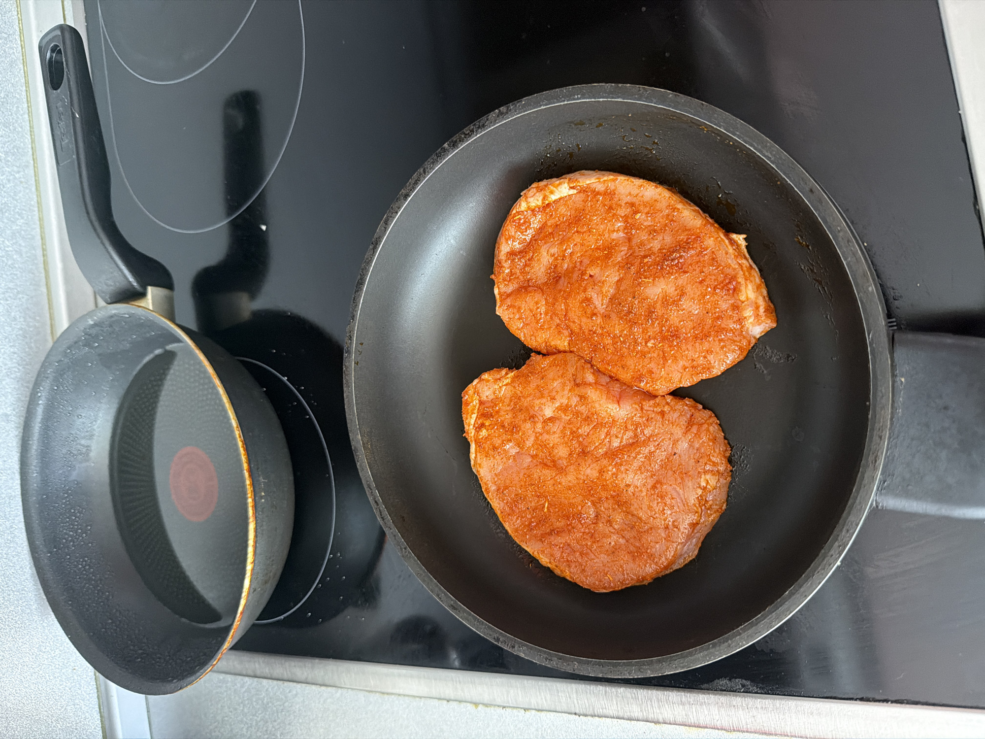Two pieces of meat, likely chicken or pork cutlets, generously coated in a reddish-orange spice rub, are centrally placed within a black frying pan. To the left, a smaller, empty black saucepan with a prominent red circular heat indicator in its center rests on the stovetop. Both pans are situated on a glossy black ceramic or induction stovetop, which features circular outlines for its burners. A light-colored wall or countertop is visible along the left and bottom edges of the frame.
The image captures a moment of food preparation, with marinated meat positioned in a pan, either awaiting or beginning to cook on the stovetop. The smaller pan suggests other ingredients may be prepared simultaneously.
Notable details include the vibrant seasoning on the meat, suggesting a flavorful dish. The larger frying pan shows traces of oil or fat around the meat. The smaller saucepan's exterior appears slightly textured or worn, and its handle has faint, illegible embossed text, possibly a brand name like "Tefal". Reflections of overhead lighting or a window are visible on the shiny stovetop and pan surfaces. The scene is set in an indoor kitchen. No specific time of day or weather conditions are discernible from the image. The provided location context is Plovdiv, Bulgaria. No people are visible.