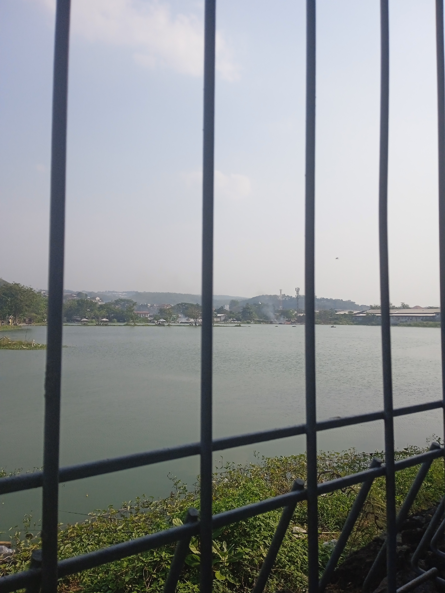 The image shows a calm lake with a distant treeline dotted with buildings and structures, including cell towers. The foreground is dominated by a metal fence, obscuring parts of the view. The water is a muted green, reflecting a hazy sky. Small boats can be seen on the water, and some smoke rises from the shore. Vegetation lines the edge of the lake. The scene appears to be a park or recreational area within or adjacent to an urban environment, possibly in Semarang, Indonesia, given the presence of what could be traditional Indonesian gazebos or huts along the shore. The overall atmosphere is serene, despite the urban elements visible in the distance.