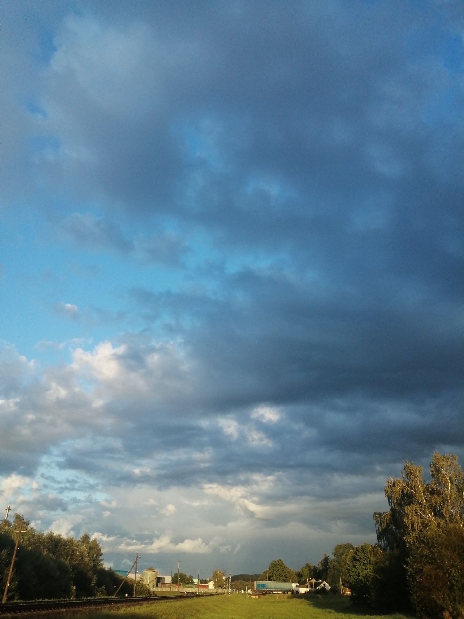 The image depicts a rural landscape with a railway track in the foreground. The sky is filled with a dramatic array of clouds, ranging from dark, stormy masses to lighter, scattered formations. To the left, a dense line of green trees borders the railway. In the middle ground, several industrial buildings with silo-like structures are visible. To the right, a truck with a blue trailer is seen on a road crossing the railway tracks, and further to the right, a large tree with sparse foliage stands. The overall lighting suggests it is late afternoon or early evening, with shafts of sunlight breaking through the clouds in places. The scene is located in the vicinity of Slonim, Belarus.
