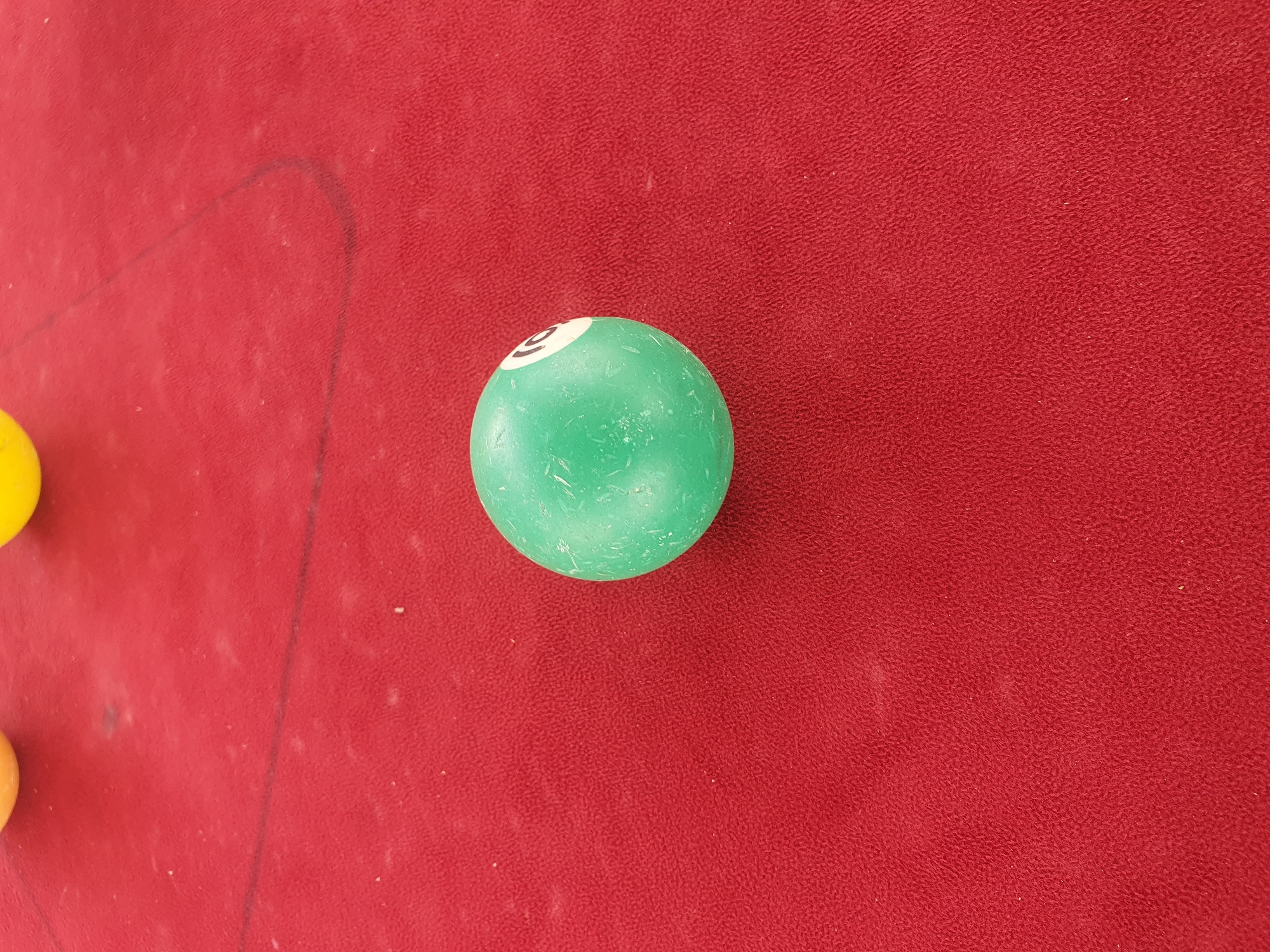 The image shows a close-up, overhead view of a green billiard ball with the number "9" printed in white on it. The ball is resting on a red surface, likely a billiard table, as indicated by the texture and color of the fabric. A faint, dark line, possibly a chalk mark or part of the table's design, curves across the red surface to the left of the ball. To the far left of the image, partially visible, are the tops of two other billiard balls, one yellow and one orange. The lighting appears to be natural and even, suggesting the photo was taken indoors or on an overcast day. The scene is static, with no discernible activity. No people are visible. The context of Lokoja, Nigeria, is provided, but there are no specific visual cues in the image to confirm this location.