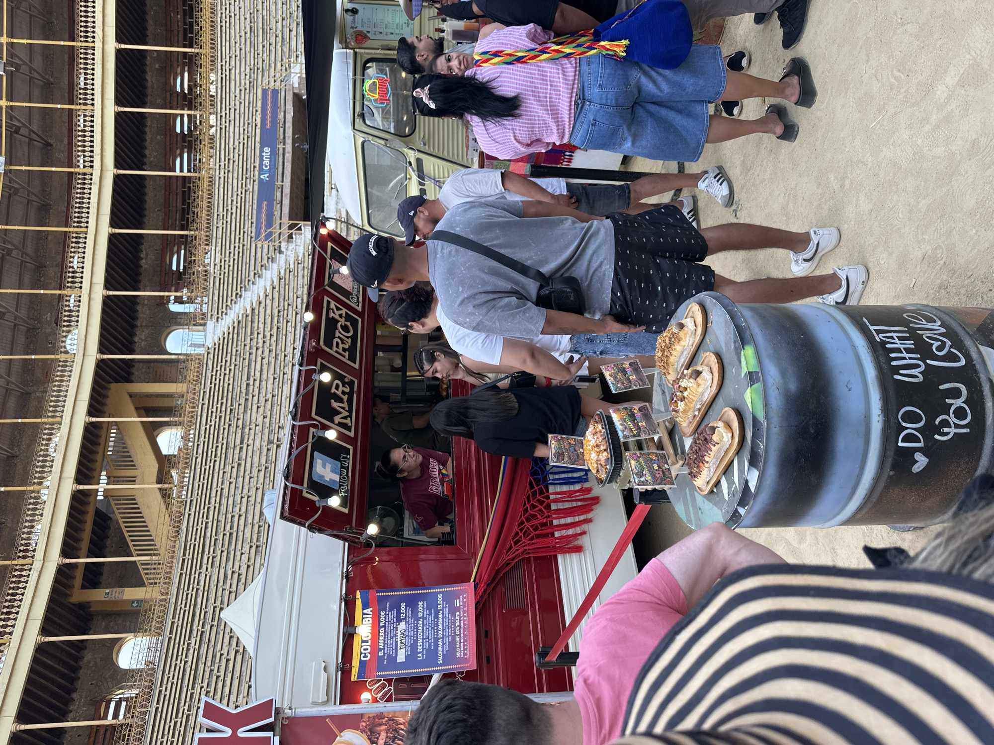 This image captures a food festival or market taking place within the Plaza de Toros de Alicante, Spain, indicated by the distinctive tiered seating, yellow-painted pillars, arches, and a "Alicante" banner in the background. The ground is sandy dirt.
Approximately 15-20 individuals, predominantly adults in casual attire, are distributed across the scene, gathering around food vendors. In the foreground, the back of a person wearing a black and tan striped hat or garment is visible. Adjacent to them, a display of food items, including several elongated pastries on wooden trays and a larger tray of what appears to be loaded fries, is arranged atop two stacked black barrels. The bottom barrel is painted with the text "DO WHAT YOU LOVE."
Centrally positioned is a red food truck displaying "MR. RICK" and "FOLLOW US!" with a Facebook logo. A "COLOMBIA" menu board lists dishes with prices in Euros, such as "EL ARRIERO: 11.00€", "TUMBAO: 12.00€", "LA DESECHADA: 12.00€", and "SALCHIPAPA COLOMBIANA: 15.00€". A hammock in Colombian flag colors (yellow, blue, red) is draped beneath the serving window. A woman in a maroon shirt is visible inside the truck, looking towards the customers. Red barrier tape is deployed in front of the truck, delineating a queuing area. A second, lighter-colored food truck with a "Tacos" sign is partially visible behind the red one.
Attendees are standing and observing the food offerings. One man in a black baseball cap and grey t-shirt with a black shoulder bag is seen from behind. To the right, a woman with long dark hair, wearing a pink plaid shirt and blue denim culottes, carries a vibrant, multi-colored woven shoulder bag. The overall environment suggests an active, open-air culinary event.