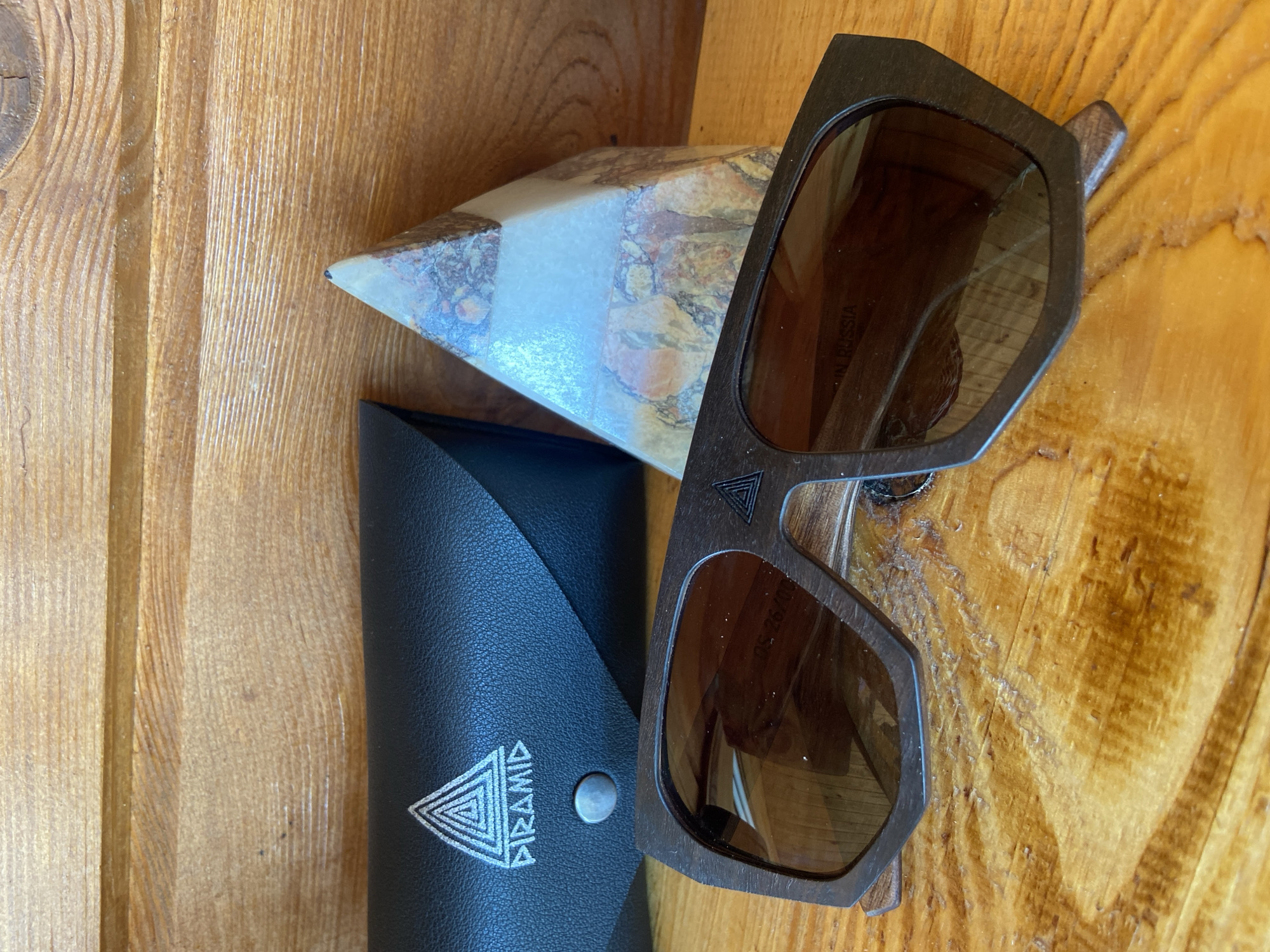 The image displays a pair of wooden sunglasses with brown lenses resting on a wooden surface. To the left of the sunglasses is a black leather case with a triangular logo and the word "PIRAMID" printed on it. To the right of the sunglasses is a layered crystal pyramid. The background consists of a wooden plank wall. The scene is lit by natural light, casting subtle shadows. The overall setting appears to be indoors, on a table with a wooden grain pattern. There are no people or animals present in the image.