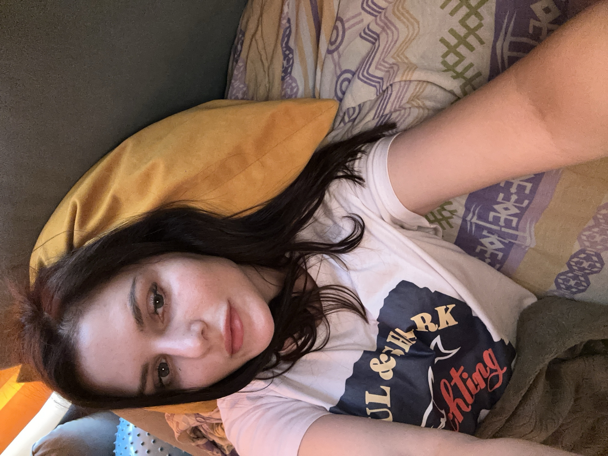 The image features a young woman lying on a bed. She has long, dark hair and is looking towards the camera with a soft expression. She is wearing a white t-shirt with a graphic design that appears to have text. Partially visible text includes "RK" in gold and "fighting" in red, with other text in dark blue. She is lying on a patterned comforter with purple and beige stripes and circular designs. A yellow pillow is positioned behind her head and shoulder. The scene appears to be indoors, with muted lighting suggesting it could be daytime or evening. The overall mood is relaxed and intimate, as the woman is captured in a casual, personal moment.