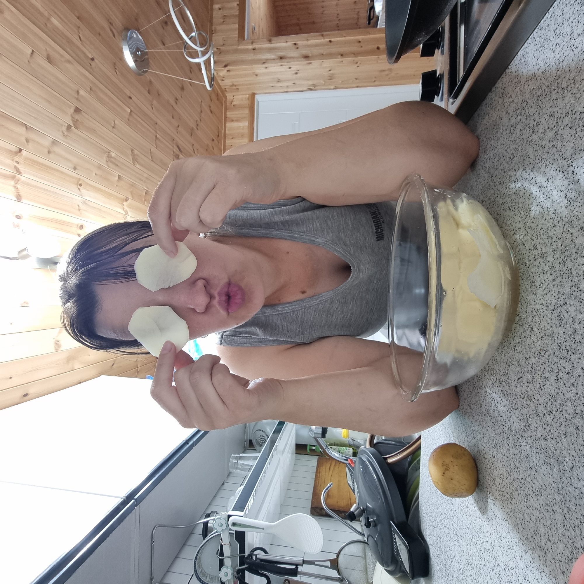 A person with dark hair, wearing a gray tank top, is in a kitchen with wooden paneling on the ceiling and walls. They are holding two thin, circular slices of potato over their closed eyes while puckering their lips. In front of them is a glass bowl filled with water and potato slices. A single whole potato sits on the counter to their left. To the right, a stovetop with a black pan is visible. The overall scene appears to be a candid, humorous moment captured in a domestic setting.