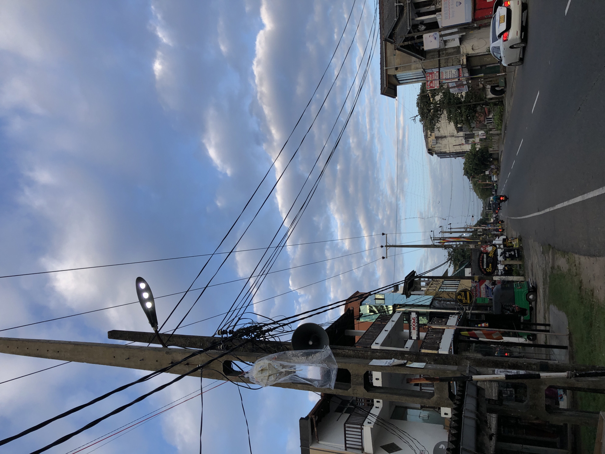 This media file depicts a street scene in Habaraduwa, Sri Lanka. The image is oriented vertically, with the sky taking up a significant portion of the frame. The sky is filled with dramatic, overcast clouds, suggesting it might be late afternoon or early evening, or a cloudy day.
The foreground is dominated by a utility pole, heavily laden with electrical wires. A street light with three lamps is attached to the pole, emitting a bright light. Below the street light, a large, dark speaker is mounted, covered in a clear plastic bag.
The midground reveals a street lined with buildings. These appear to be shops and residences, with multiple stories and balconies. Various signs are visible, some with text in a language that is likely Sinhala, given the location. One sign indicates "ROOMS," suggesting accommodation. A white car is parked on the side of the road near one of the buildings. Further down the street, a green tuk-tuk is visible, along with traffic signals showing red lights, indicating a stopped flow of traffic.
The background shows more of the street and buildings, stretching into the distance. The road itself is paved and appears to be a main thoroughfare. The overall scene conveys a sense of a bustling, everyday street in a town in Sri Lanka, with the weather adding a slightly dramatic atmosphere. There are no people clearly visible in the frame, though their presence is implied by the activity on the street.