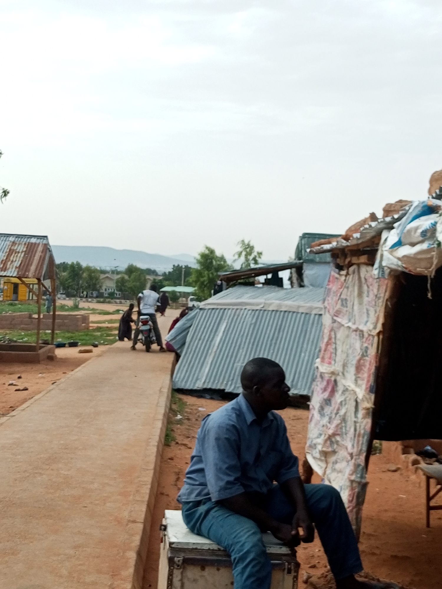 The image depicts an outdoor scene in the city of Gombe, Nigeria. In the foreground, a Black man wearing a blue collared shirt and blue jeans is seated on a white chest. He is looking to his left. Behind him, to his left, a corrugated metal roofed structure is visible. To the right of the seated man, another makeshift structure with a corrugated metal roof and walls made of what appears to be sacks and sheeting is present. A dirt path runs horizontally across the mid-ground of the image, with a concrete curb. On the path, a man is standing next to a motorcycle, with another person, possibly a child, behind him. Further in the background, there are trees, more buildings with metal roofs, and a distant hillside. The sky is overcast. The ground in the foreground and mid-ground is composed of reddish-brown earth.