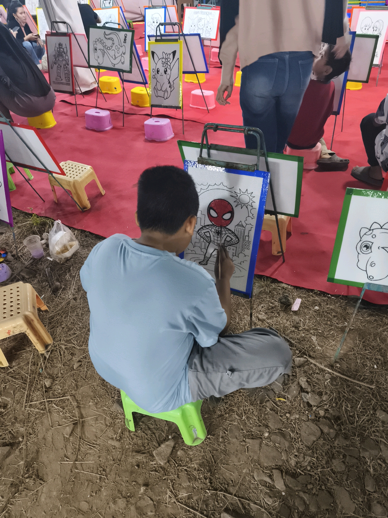 In Palembang, Indonesia, a young boy sits on a green plastic stool on a dirt and dried grass ground, facing away from the camera. He is wearing a light blue t-shirt and gray pants, and is actively coloring a drawing of Spider-Man on a canvas. The canvas is mounted on a blue easel. In the background, there is a red carpeted area with more easels displaying various cartoon character drawings, such as Pikachu and a robot. Several small yellow and pink plastic stools are scattered around the red carpet. Other people are visible in the background, including a woman with dark hair and a person wearing a red shirt. The scene appears to be an outdoor activity or event where children can engage in coloring.