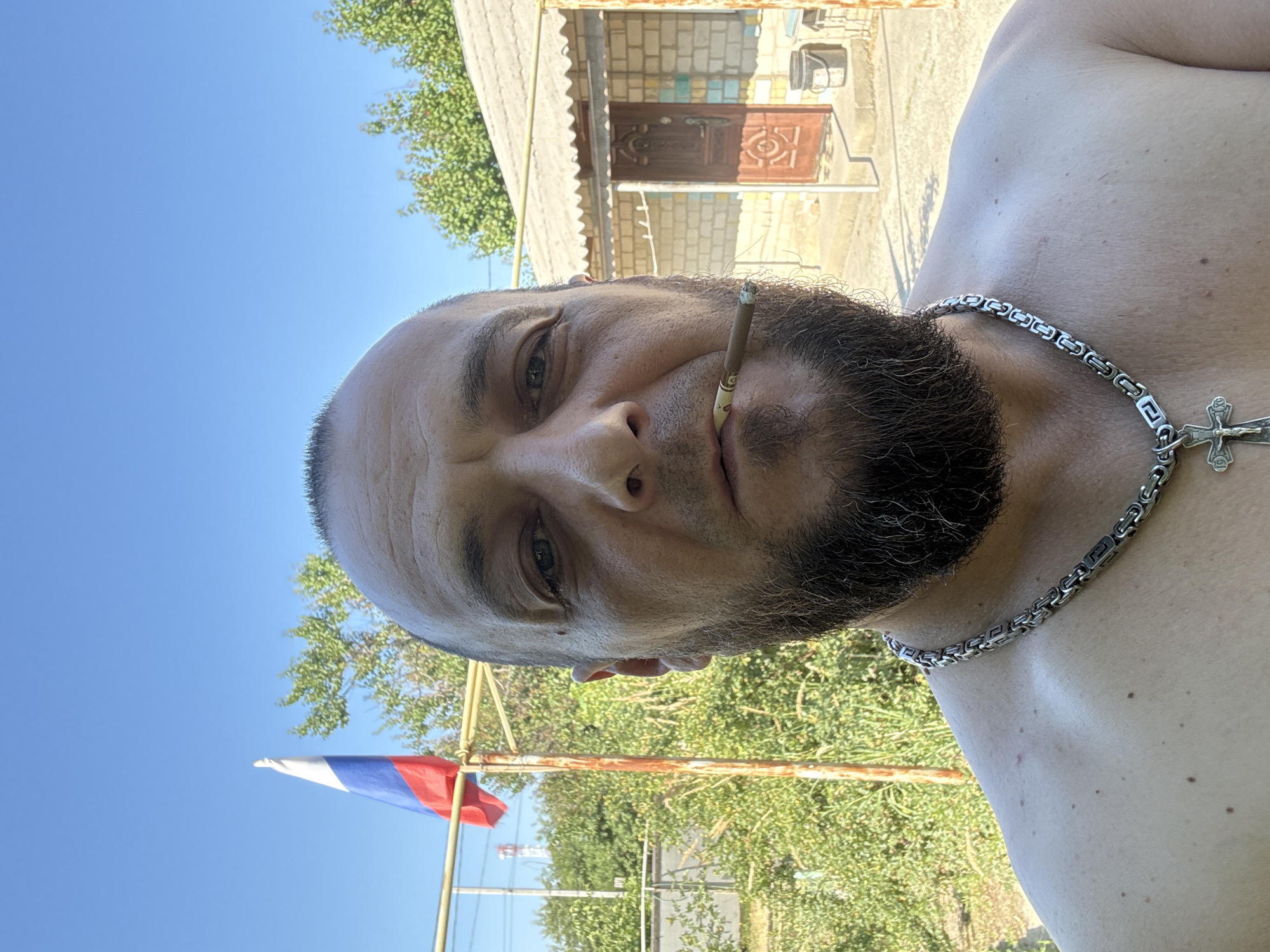 A man with a beard and a bald head is shown from the chest up, facing forward. He has a cigarette in his mouth and is wearing a silver chain with a cross pendant. In the background, a Russian flag flies from a pole, and a brick building with a brown door is visible. The scene appears to be outdoors, with greenery and a clear blue sky. The man's bare shoulders are visible. This scene is located in the city of Zhirnov, Russia.