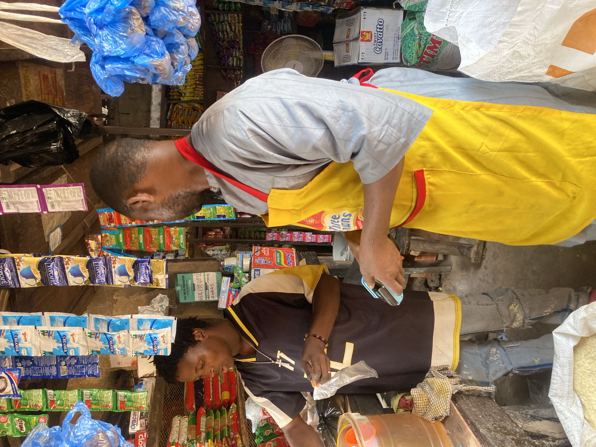 This image depicts the interior of a general goods shop or market stall in Rido, Nigeria. Two individuals are present, engaged in commercial activities. On the left, a person with dark skin and short hair, wearing a black and yellow short-sleeved top with the number "77" and a cross pendant, distressed blue jeans, and a beaded bracelet, is handling a small clear plastic bag, appearing to package an item from a transparent container on a wooden counter. To their right, a second person with dark skin and short dark hair, wearing a light grey long-sleeved garment and a yellow apron with red trim, holds a blue and black mobile point-of-sale (POS) device in their right hand.
The environment is densely packed with merchandise. Above the individuals, various packaged goods hang from the ceiling and are displayed on shelves. These include sachets of Milo, Creamer, Topcafe, and Onga seasonings. A sign with "PAY WITH TRANSFER," "O-Pay," and account details including "ABDULLAHI ABBAS" is taped to a wall in the background. Shelves behind the individuals hold stacks of packaged foods like pasta and other items. A white electric fan is visible on a shelf to the right. Numerous blue plastic bags, some containing goods, hang from the ceiling on the right side. A large white woven sack filled with a granular substance, likely rice, is situated on the floor in the foreground on the left. A cardboard box labeled "CROWN TWIST Cavatto" is visible near the fan. The floor appears to be concrete, and the walls show signs of wear.