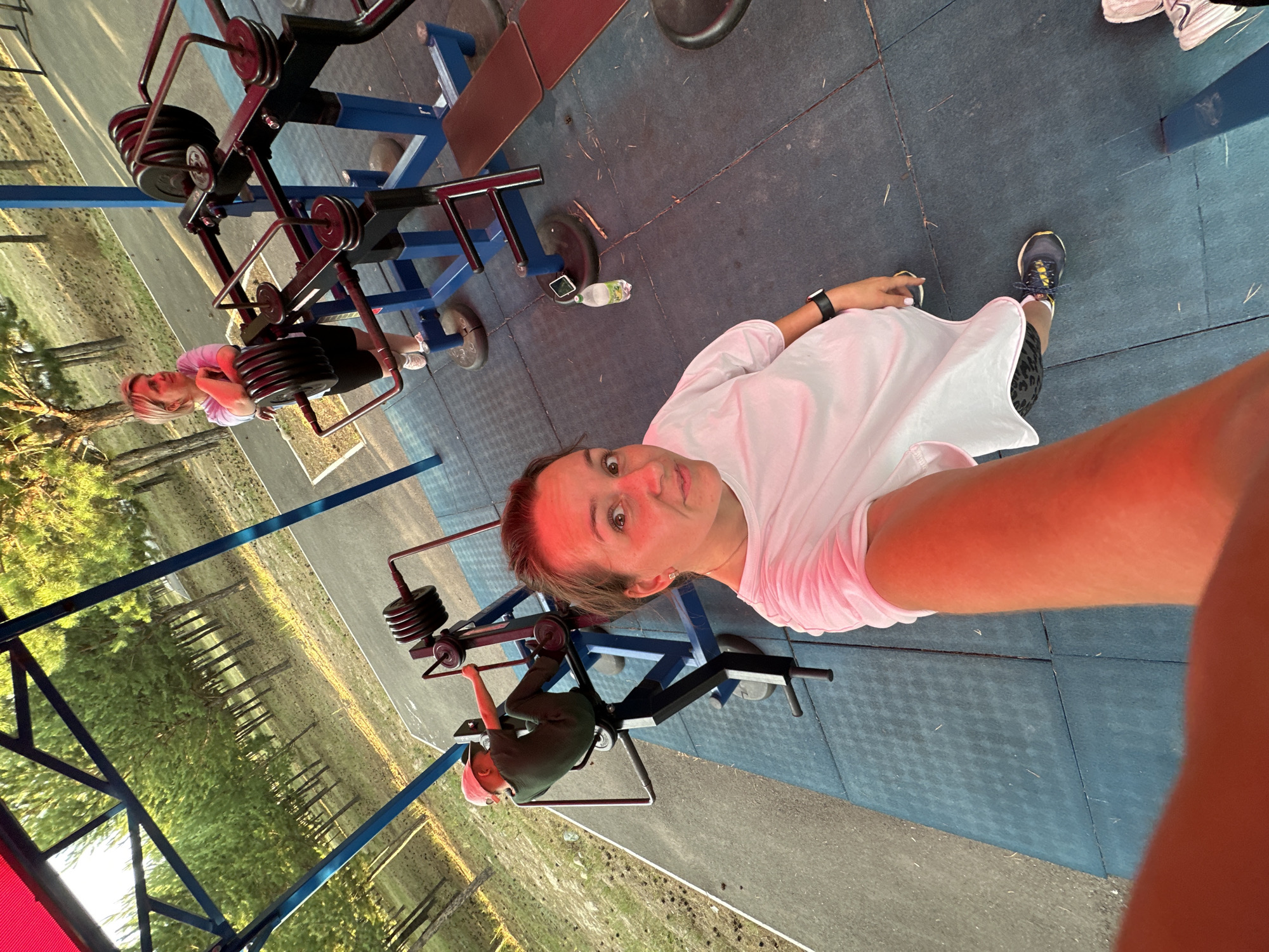 The image shows two people at an outdoor gym in Bely Yar, Russia. The primary subject is a woman in the foreground, wearing a white t-shirt and dark shorts, taking a selfie. She has her arm extended, holding the camera. Her expression is direct and engaging. To her left, a child is seated on a weight machine, wearing a green t-shirt and a pink cap. The child is facing away from the camera and appears to be operating the machine.
The setting is an outdoor fitness area with blue and red gym equipment. Weight plates are visible on the machines. The ground is covered with dark blue rubber matting. In the background, there is greenery with scattered trees, suggesting a park or natural area. The lighting indicates it is daytime, possibly late afternoon given the warm tones and shadows. The weather appears to be clear. No visible text is present in the image. The overall mood is casual and active, with the individuals engaged in a workout.