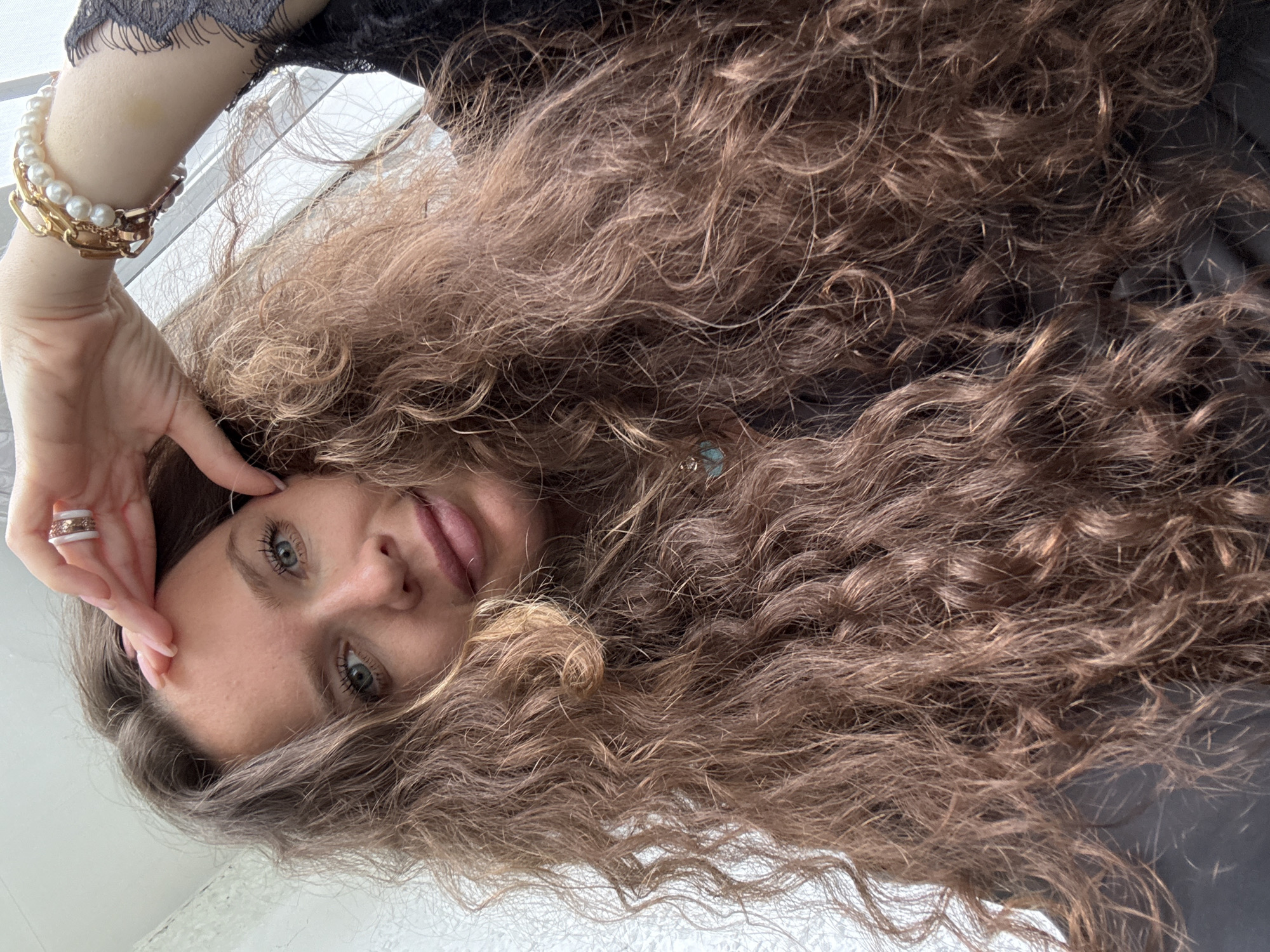 The image shows a close-up, selfie-style shot of a woman with long, curly, brown hair. She has fair skin, light blue eyes, and is wearing light makeup. Her right hand is raised, touching her forehead, and she is wearing a ring on her index finger and a bracelet on her wrist. She is wearing a black top with lace details. The background appears to be a lightly textured wall. There is no information about the location in Turinsk, Russia.
