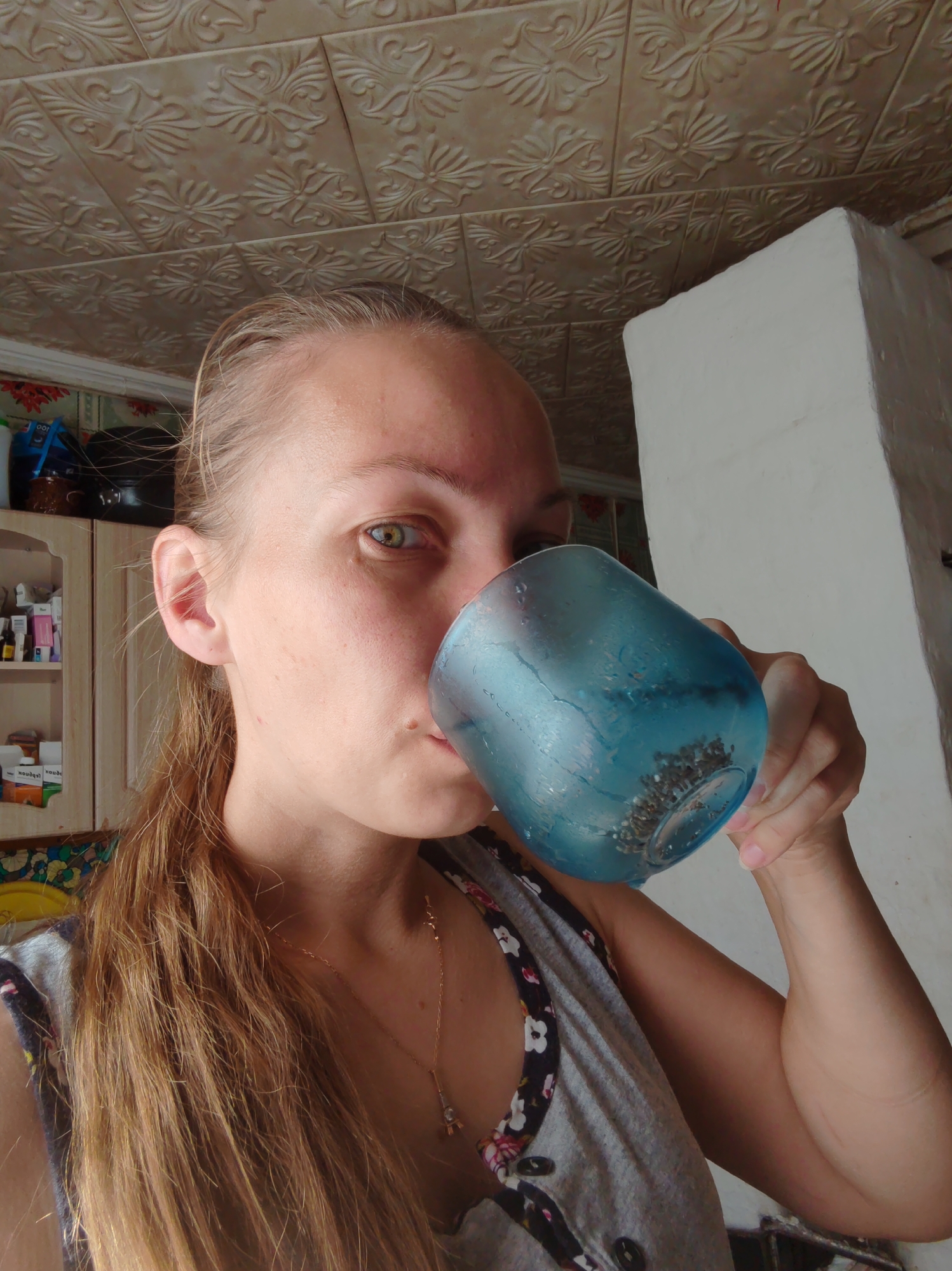A close-up, over-the-shoulder shot shows a young Caucasian woman with long, light brown hair drinking from a blue glass. The woman is positioned in the foreground, her face angled towards the viewer as she sips from the glass. She is wearing a grey tank top with a floral trim. Behind her, a white plaster structure, possibly a stove, is visible, along with a patterned ceiling and a cabinet with various items on its shelves. The scene appears to be indoors, in what could be a kitchen or living area, in an urban setting. The lighting is dim, suggesting it may be daytime with natural light filtering in. The overall atmosphere is casual and domestic.