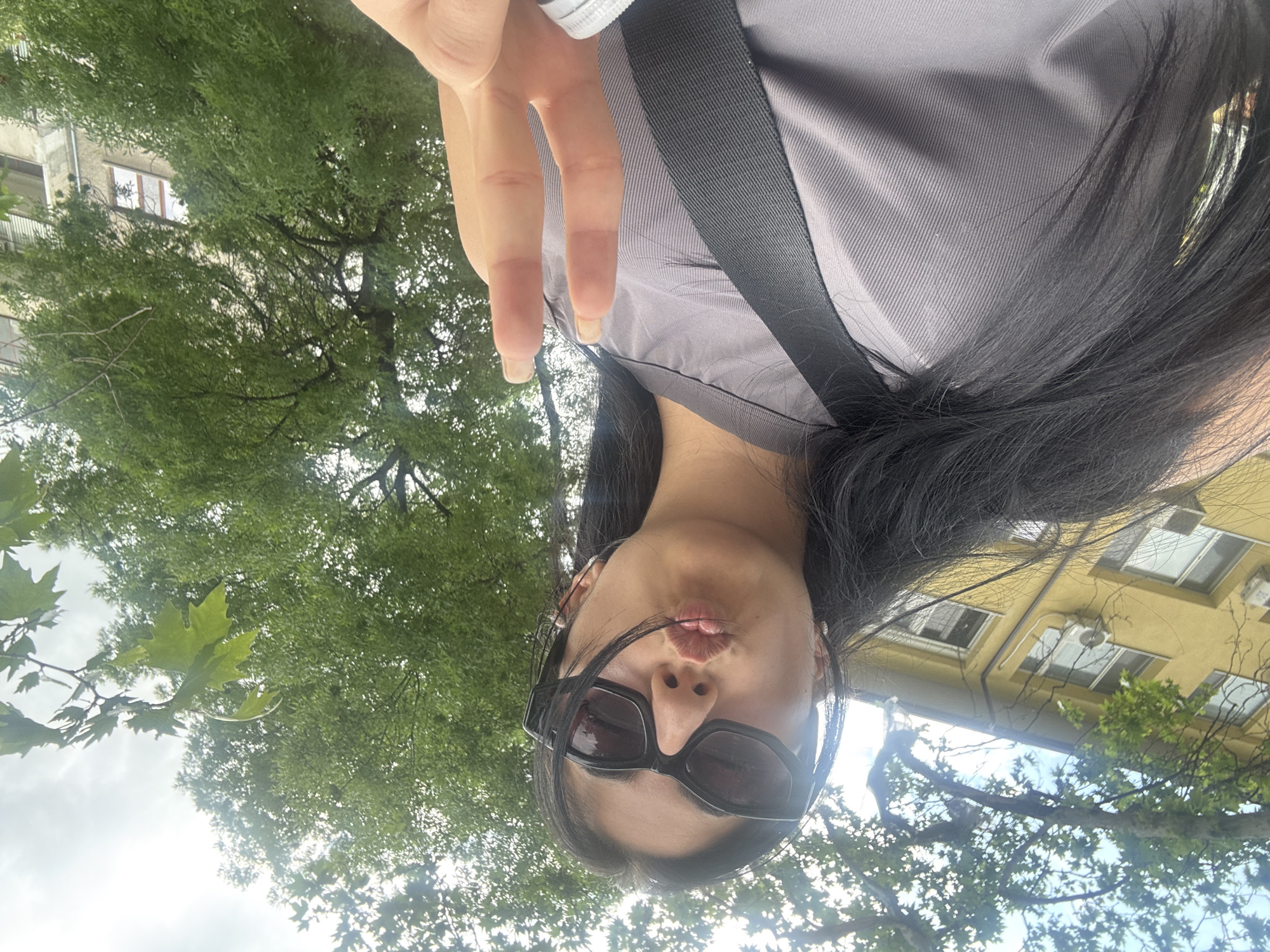 A woman with long, black hair, wearing dark sunglasses and a gray top with a black strap, is taking a selfie. She is making a kissing face and holding up her fingers in a peace sign. Behind her is a green tree and the facades of buildings. The location appears to be in an urban area in Plovdiv, Bulgaria.