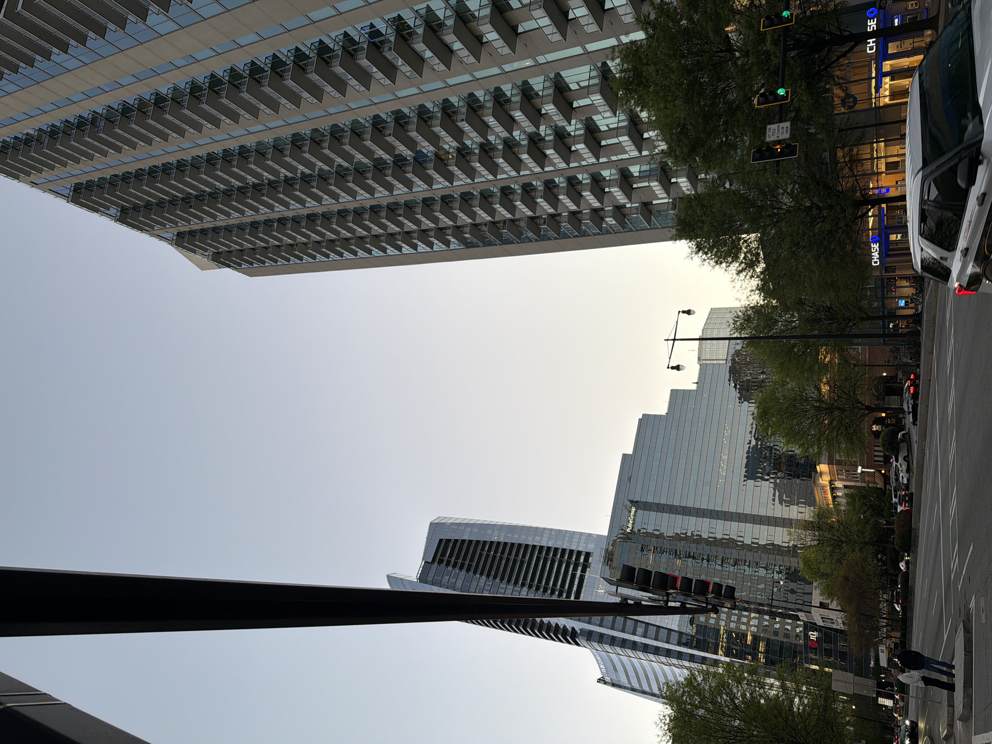 The image shows a street scene in Atlanta, United States. The view is looking upwards, capturing several modern skyscrapers against a light sky, likely during twilight. A tall residential building on the left side of the frame features numerous balconies. On the right, a street is visible with cars, trees, and buildings. Chase bank storefront is prominently displayed. A traffic signal is illuminated in green. Some people can be seen walking on the sidewalk.
