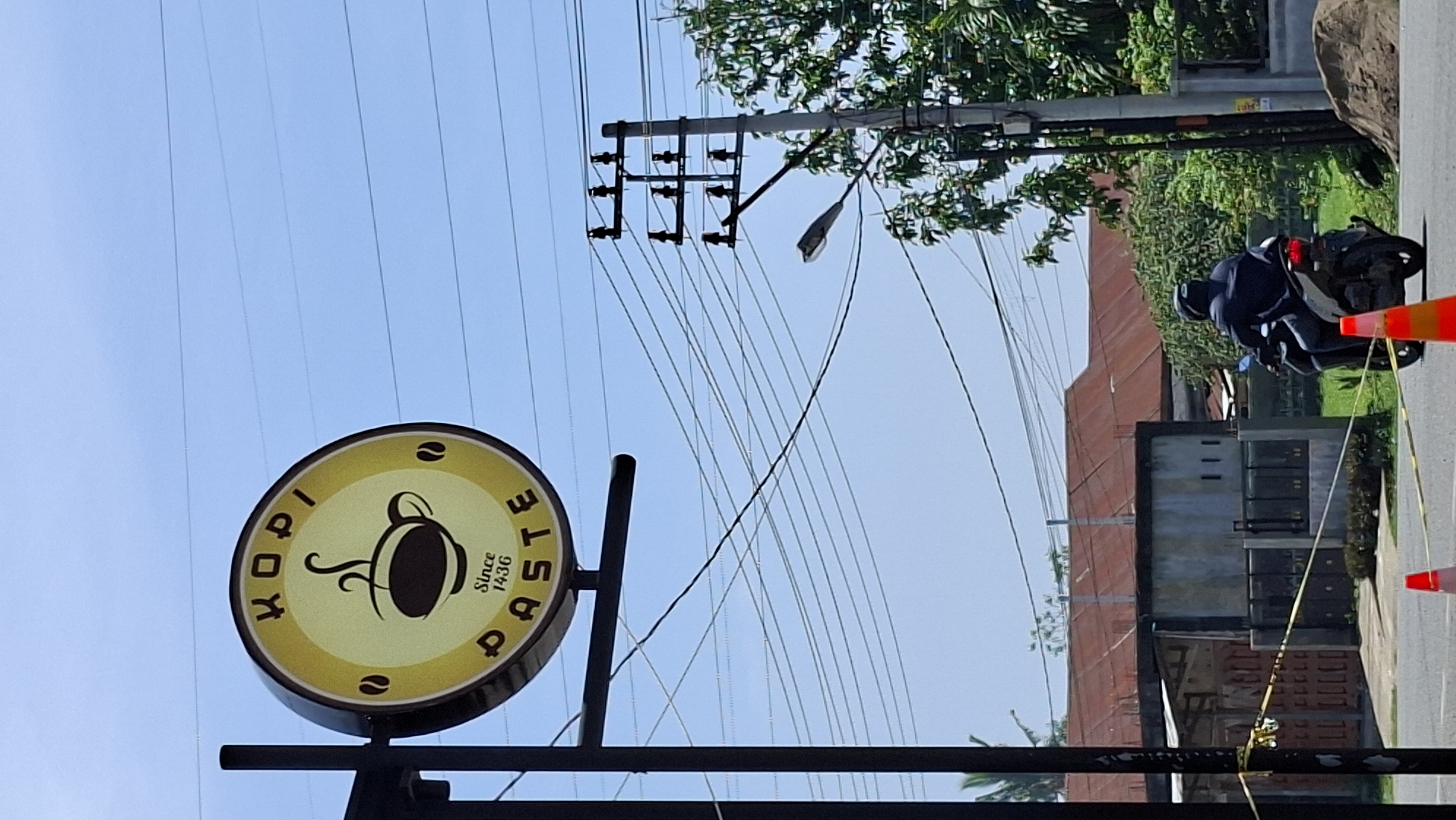 This is a vertical, eye-level shot taken outdoors, likely in a semi-urban area of Lhokseumawe, Indonesia. The primary focus is a circular sign for "KOPI PASTE Since 1936," with a stylized coffee cup and steam graphic at its center. The sign is mounted on a black pole. In the foreground, a person wearing a dark jacket and helmet is riding a dark-colored motorcycle away from the camera. The motorcycle has its brake light illuminated. Orange traffic cones are visible on the road. Behind the motorcycle and sign, there are buildings with red roofs, a concrete wall, and lush green trees. Overhead, a clear blue sky is crisscrossed with numerous electrical wires supported by utility poles. The overall scene suggests a daytime view of a commercial establishment and street activity.
