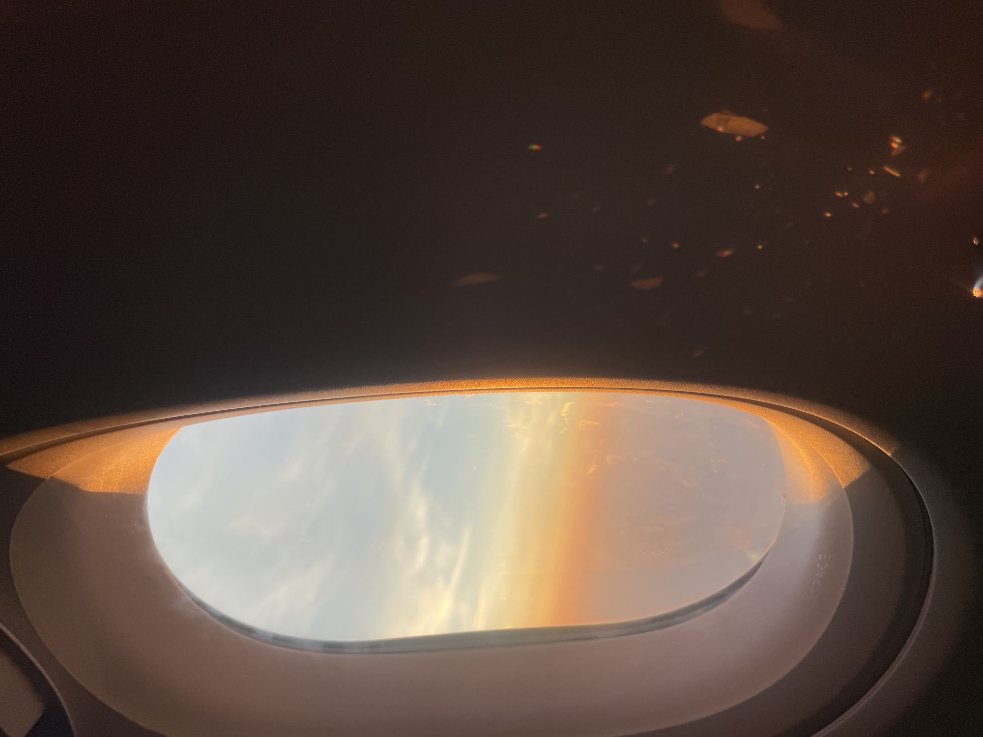 The image shows the interior of an airplane window with a view of a sunset. The window frame is visible on the left side of the image, with the setting sun casting a warm glow on the clouds and the window. The sky transitions from a bright orange and pink near the horizon to a pale blue at the top. The right side of the image is dark, with reflections or light flares visible, suggesting the interior of the airplane cabin is dimly lit. There is text embossed on the window frame that appears to say "AEROSPACE". The scene is captured from the perspective of a passenger seated inside the aircraft. There are no people or specific city locations, including St Albans, United Kingdom, visible in this image.