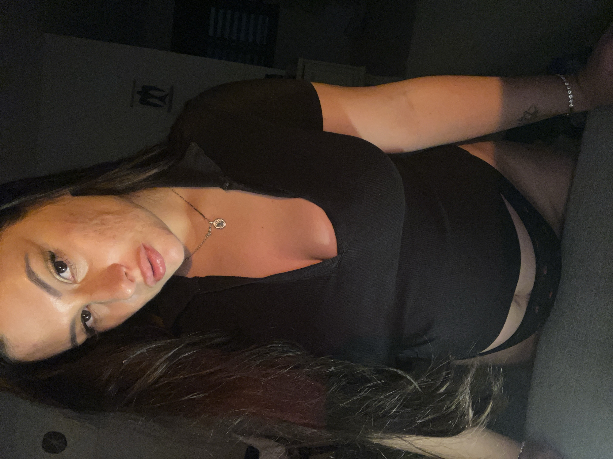 An indoor photograph, taken in Kiryat Bialik, Israel, depicts an adult female subject from the chest to the upper thighs in a dimly lit environment. The subject has long, dark hair, a medium skin tone, and is looking directly towards the camera. She is wearing a black, short-sleeved, ribbed V-neck top and black underwear with a subtle pattern, visible just below the top. A silver-colored chain necklace with a circular pendant is around her neck, and a silver-colored bracelet with multiple clear stones is on her right wrist. A dark-inked tattoo is visible on her right forearm. The background is a dark wall. To the left, behind the subject's head, a circular object with a radiating pattern is faintly visible. To the right, behind her shoulder, a distinct, dark, stylized silhouette resembling an eagle or bird of prey is present on the wall. Horizontal blinds and a light-colored piece of furniture are partially discernible in the far right background. The immediate foreground consists of a dark, textured surface, possibly fabric.