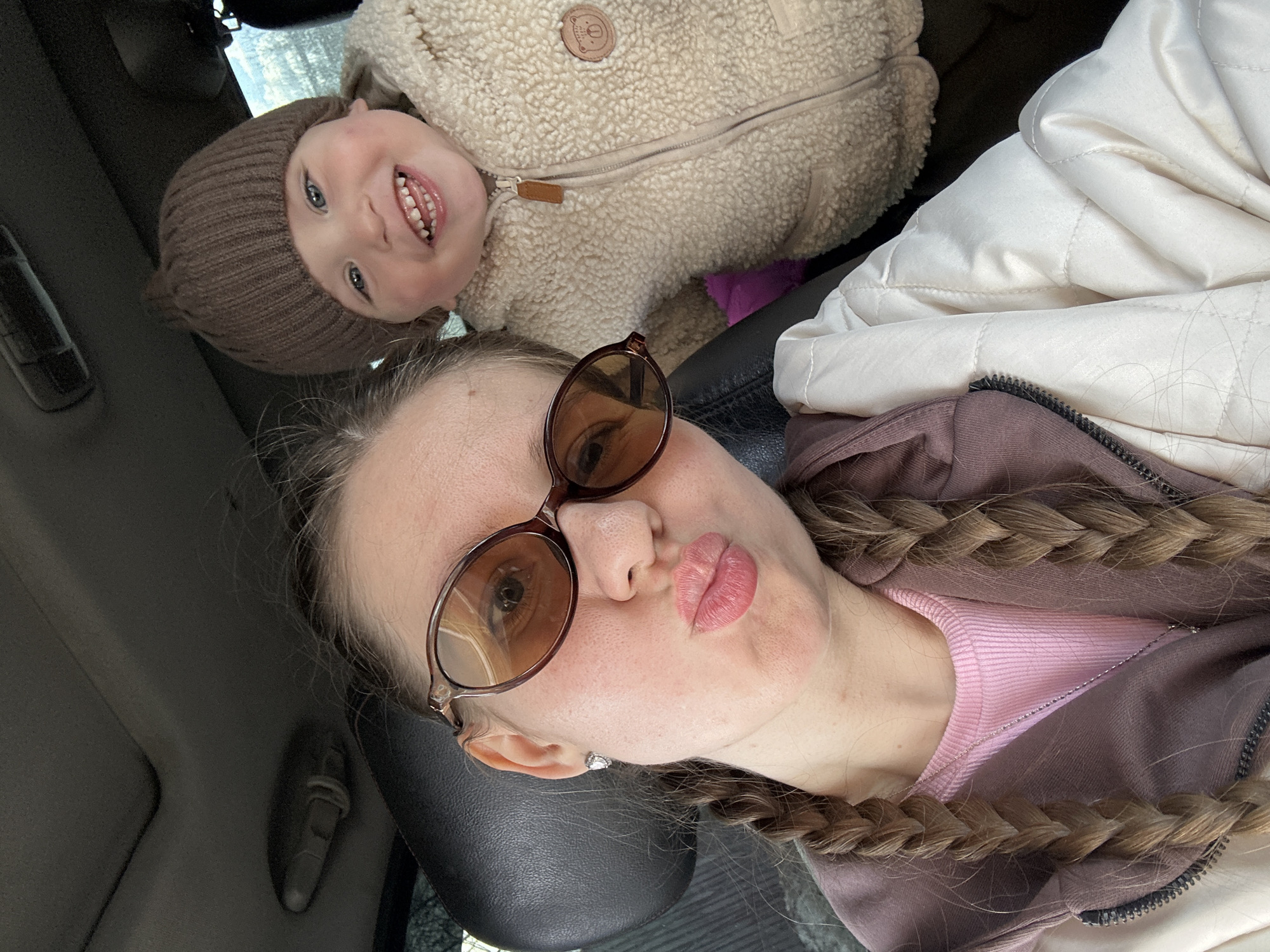 The image shows a selfie taken inside a car. A young woman with brown hair styled in braids, wearing sunglasses, a pink top, and a brown hoodie under a white jacket, is positioned in the foreground. She is puckering her lips towards the camera. Above her is a small child with a brown knit hat and a light brown fleece jacket. The child is smiling and facing the camera. The car interior is visible in the background. The location in the city of Klin, Russia, cannot be determined from the image.