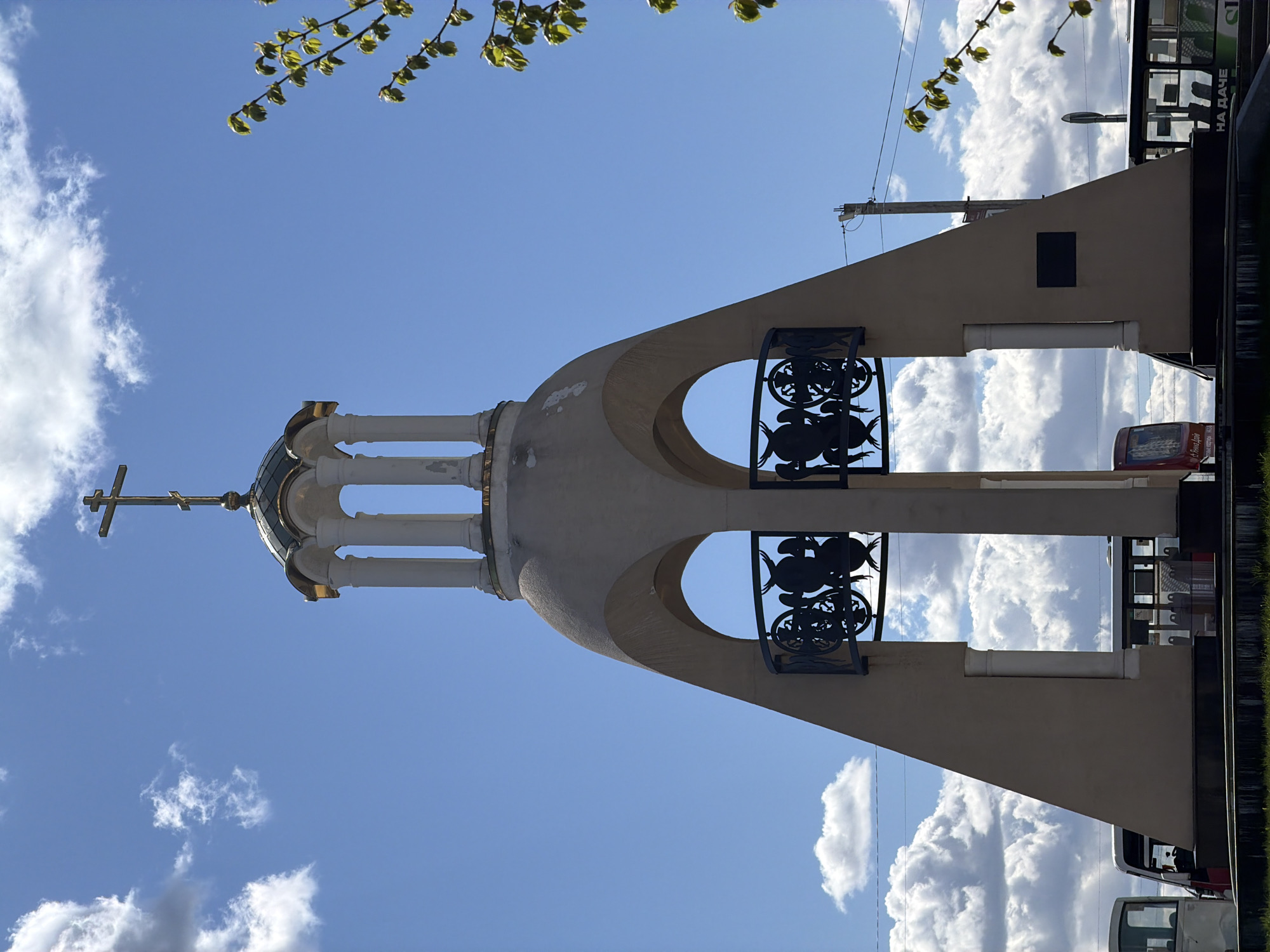 The image shows a vertical shot of a religious structure, possibly a tower or a chapel, in Koltushi, Russia. The structure is painted in light beige and features architectural elements such as arches, decorative metal railings, and a golden dome with a cross on top. A bright blue sky filled with fluffy white clouds provides the backdrop. Some green leaves are visible in the upper part of the frame. A building or structure is situated in the background, with some vehicles present.