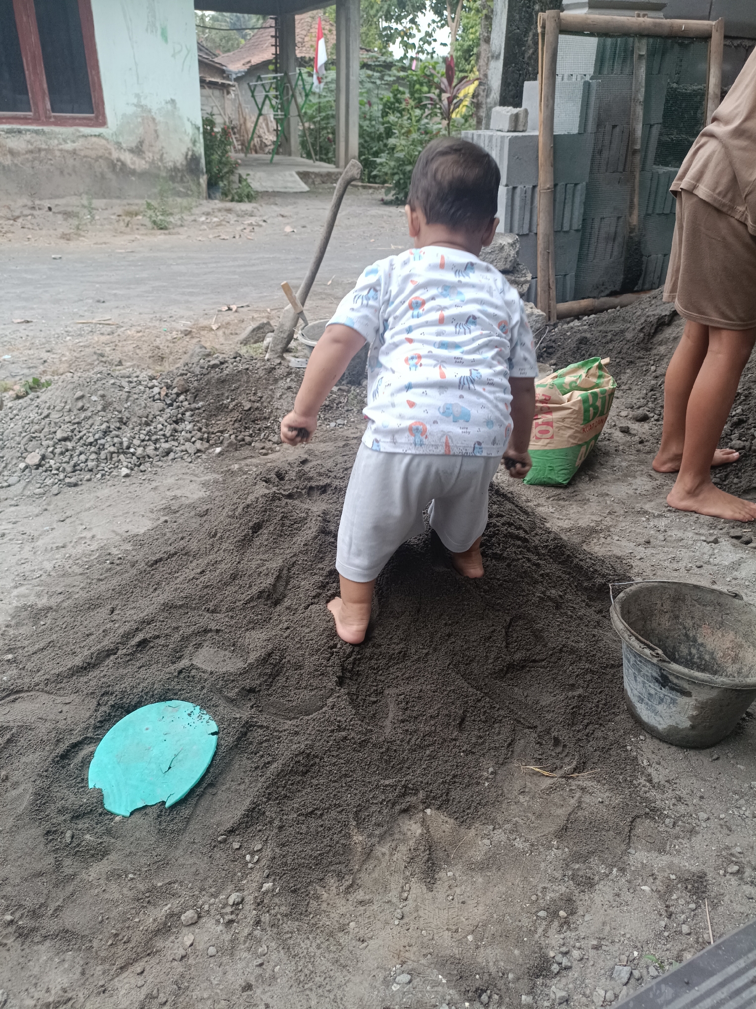 In a residential area of Sumberadi, Indonesia, a young child, viewed from the back, stands on a pile of dark sand. The child is wearing a white t-shirt with a colorful animal print and light-colored pants. The child's right hand is slightly raised, holding a small amount of sand. A teal-colored plastic lid is visible in the sand to the child's left. To the right of the child, a person's legs are visible, with the person standing barefoot. Behind the child and person, a green and yellow bag of construction material is partially visible. A weathered gray bucket is situated to the right of the sand pile. The background shows a simple building with a window, a wooden structure with mesh, and a stack of gray concrete blocks. The ground is covered with sand, gravel, and scattered debris, suggesting an outdoor construction or renovation site.