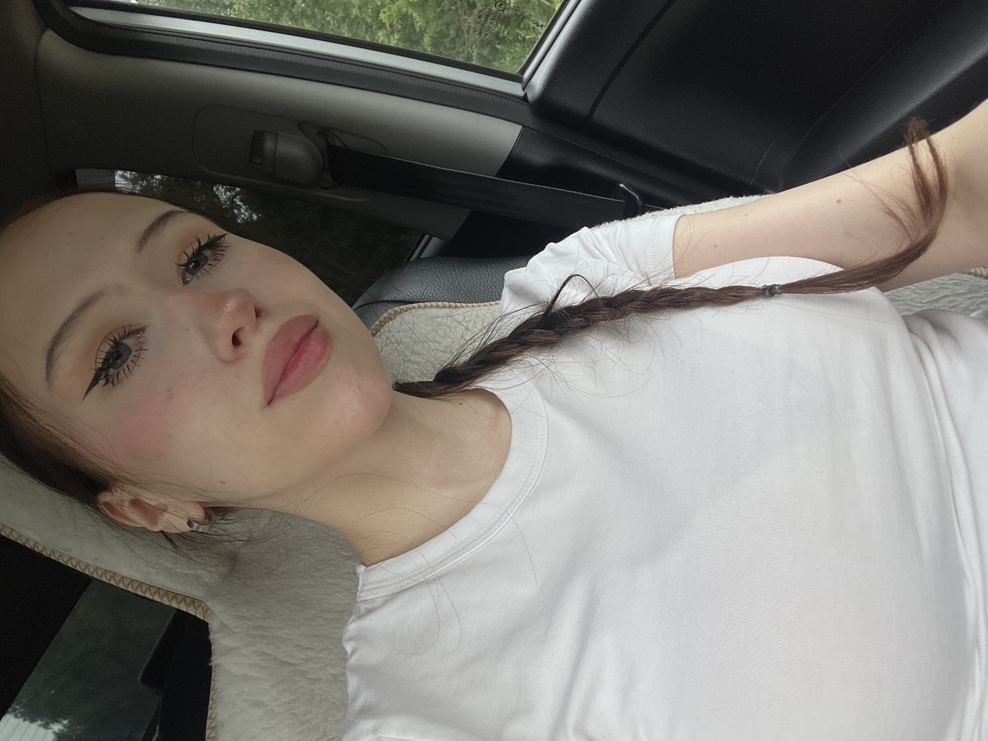A young woman with a plait of brown hair is sitting in the passenger seat of a vehicle. She is wearing a white t-shirt. Her eye makeup is dramatic with winged eyeliner and long lashes. She has blush on her cheeks and pink lipstick. The interior of the car is visible, including a seatbelt and a textured seat cover. Outside the window, green trees and foliage suggest a rural or suburban setting. There is no indication of the specific location within Gavrilovka, Russia, in the image itself.