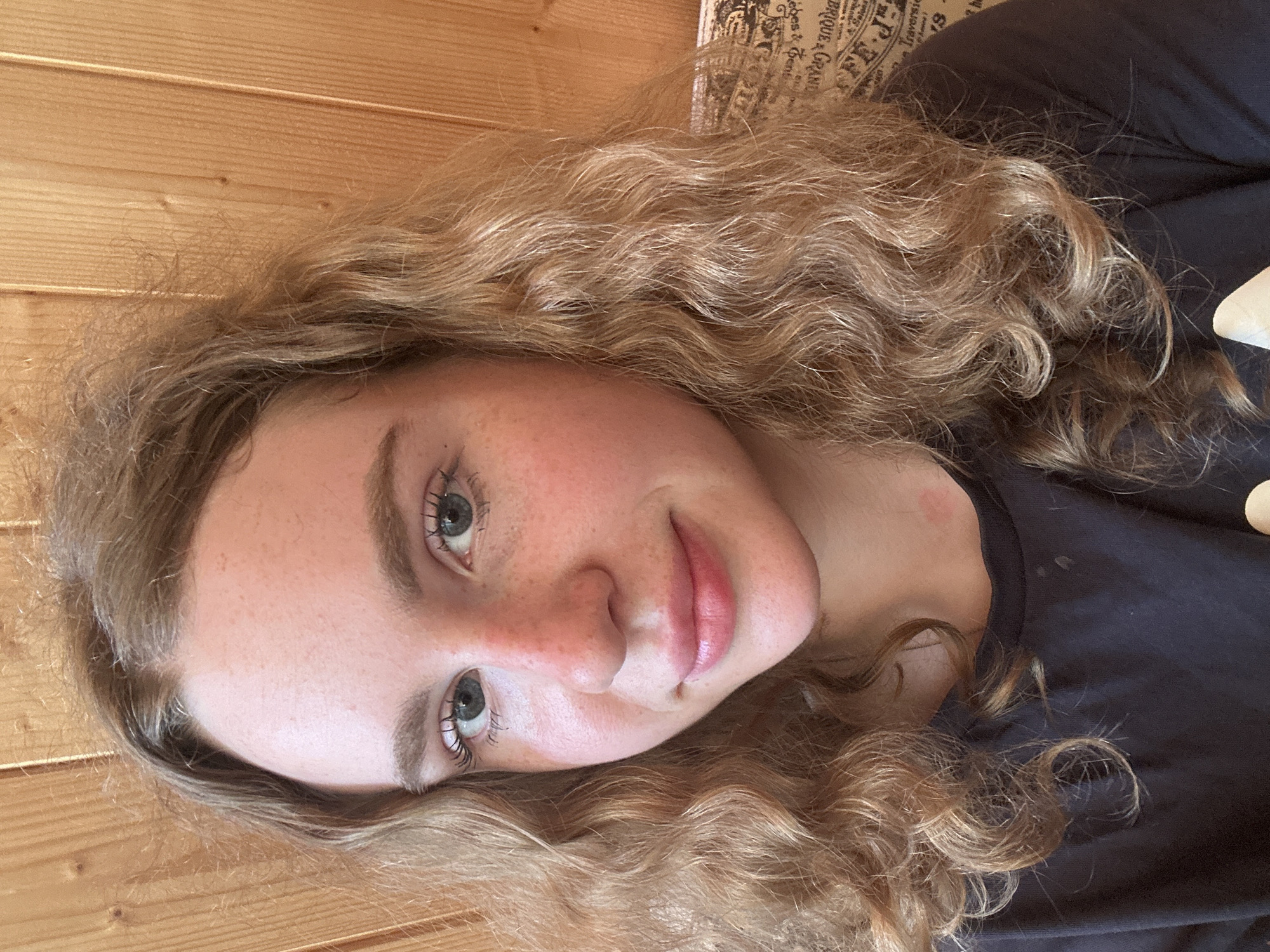 A young Caucasian female with light brown, wavy hair is positioned in the center of the frame, facing the camera. She has light skin with freckles scattered across her nose and cheeks. Her eyes are blue, and she has on mascara and lip gloss. She is wearing a black t-shirt. The background consists of vertical wooden planks, suggesting an indoor setting with a rustic aesthetic. The lighting appears natural, casting subtle shadows and highlighting the texture of the wood and her hair. There is a small red mark on her neck. The overall scene is a close-up portrait of the individual. There is no indication of the city of Vasilyevo, Russia in the image.