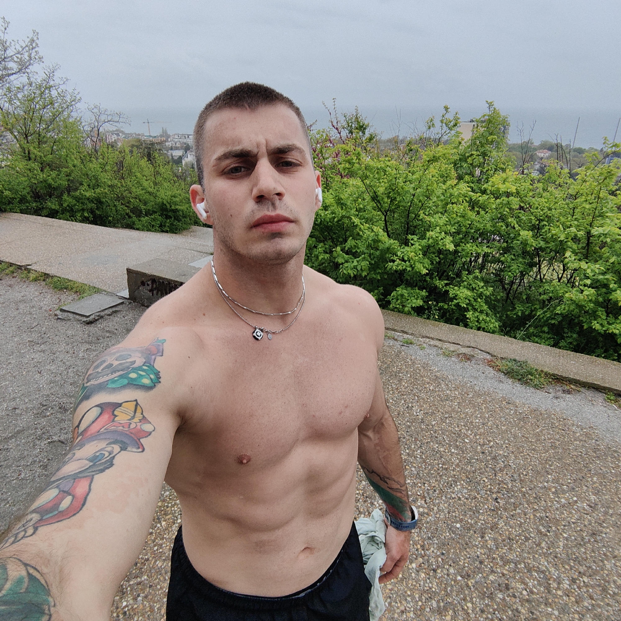 This media file captures an outdoor selfie of a shirtless man, likely taken from an elevated position overlooking a coastal city. The man, with a short buzz cut, gazes directly at the camera with a neutral expression. He wears white wireless earbuds in both ears and two silver-toned necklaces, one featuring a small black and white pendant. His muscular torso and defined abdominal muscles are visible, with hints of moisture or sweat on his skin.
He has several colorful tattoos: a stylized skull with green elements and a cartoon character resembling Donald Duck on his left arm, and green and red striped elements on his right forearm. He is dressed in black athletic shorts and wears a blue wristwatch on his right wrist, holding a light-colored cloth in his left hand.
The background features lush green trees and bushes that partially obscure a distant cityscape, including buildings and a visible crane. Beyond the city, a vast body of water, consistent with the Black Sea given the Varna context, stretches to the horizon under a heavily overcast, cloudy sky. The foreground consists of a concrete path and a gravelly surface. The overall scene suggests an outdoor workout or run on a cool, possibly damp, day. No visible text is present in the image.
